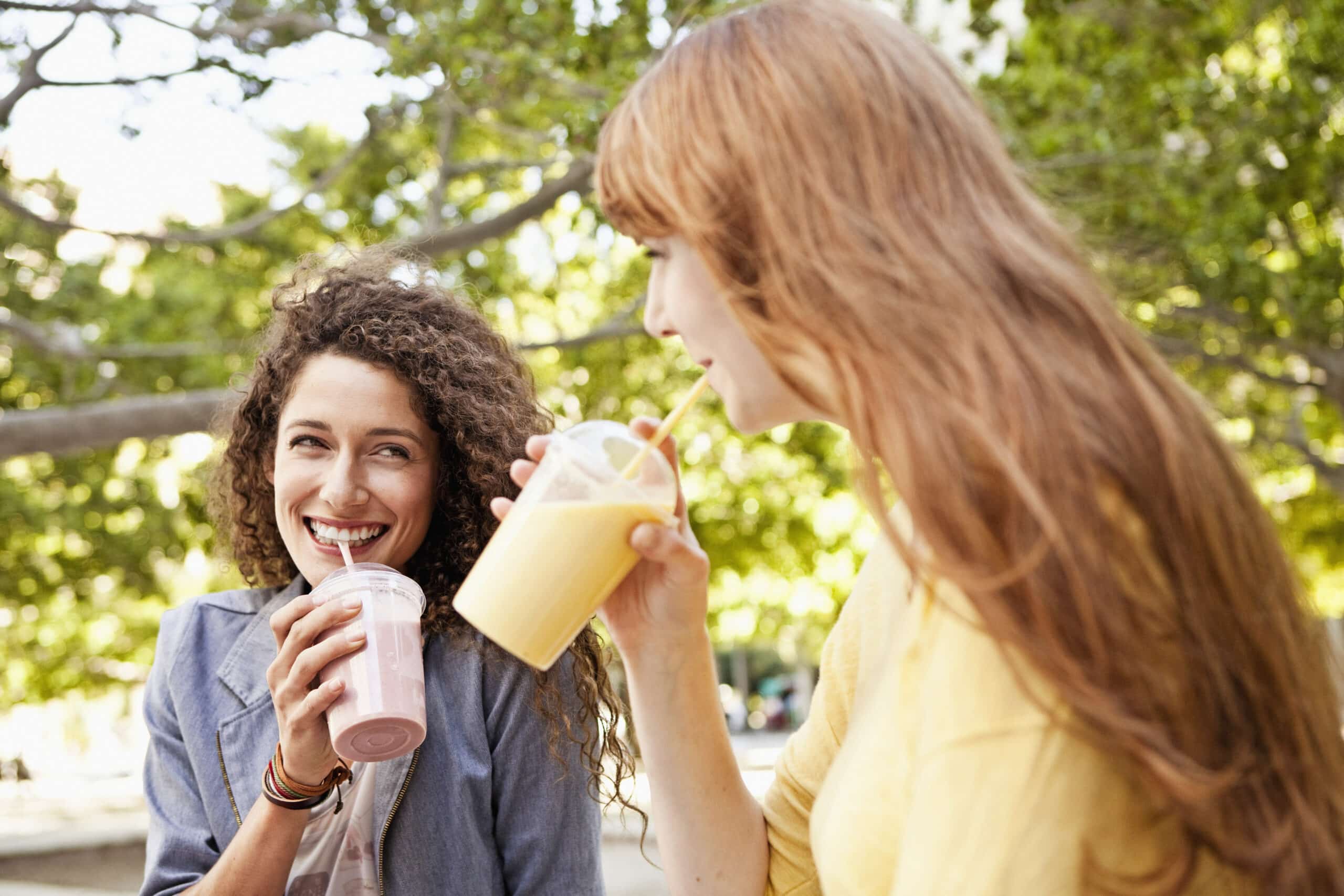 Zwei Frauen sitzen draußen und genießen Smoothies. Eine hat lockiges Haar und hält einen rosa Smoothie in der Hand, während sie die andere Frau mit langem, glattem Haar anlächelt, die an einem orangefarbenen Smoothie nippt. Sie sind von Bäumen und Sonnenlicht umgeben, was eine entspannte Atmosphäre suggeriert. © Fotografie Tomas Rodriguez