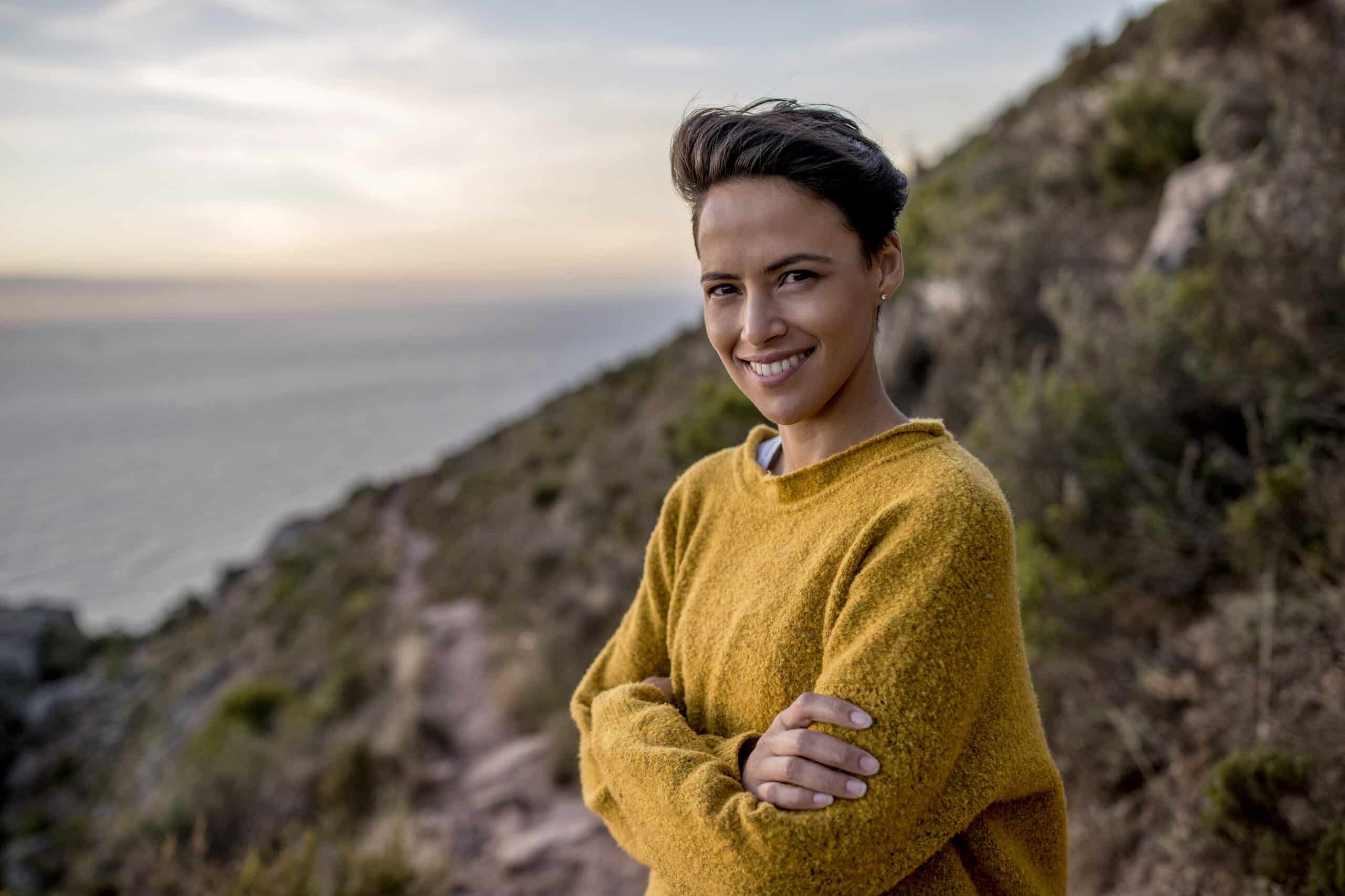 Eine lächelnde Frau in einem gelben Pullover steht bei Sonnenuntergang mit verschränkten Armen und heiterem Gesichtsausdruck auf einem Hügel mit Blick auf das Meer. © Fotografie Tomas Rodriguez