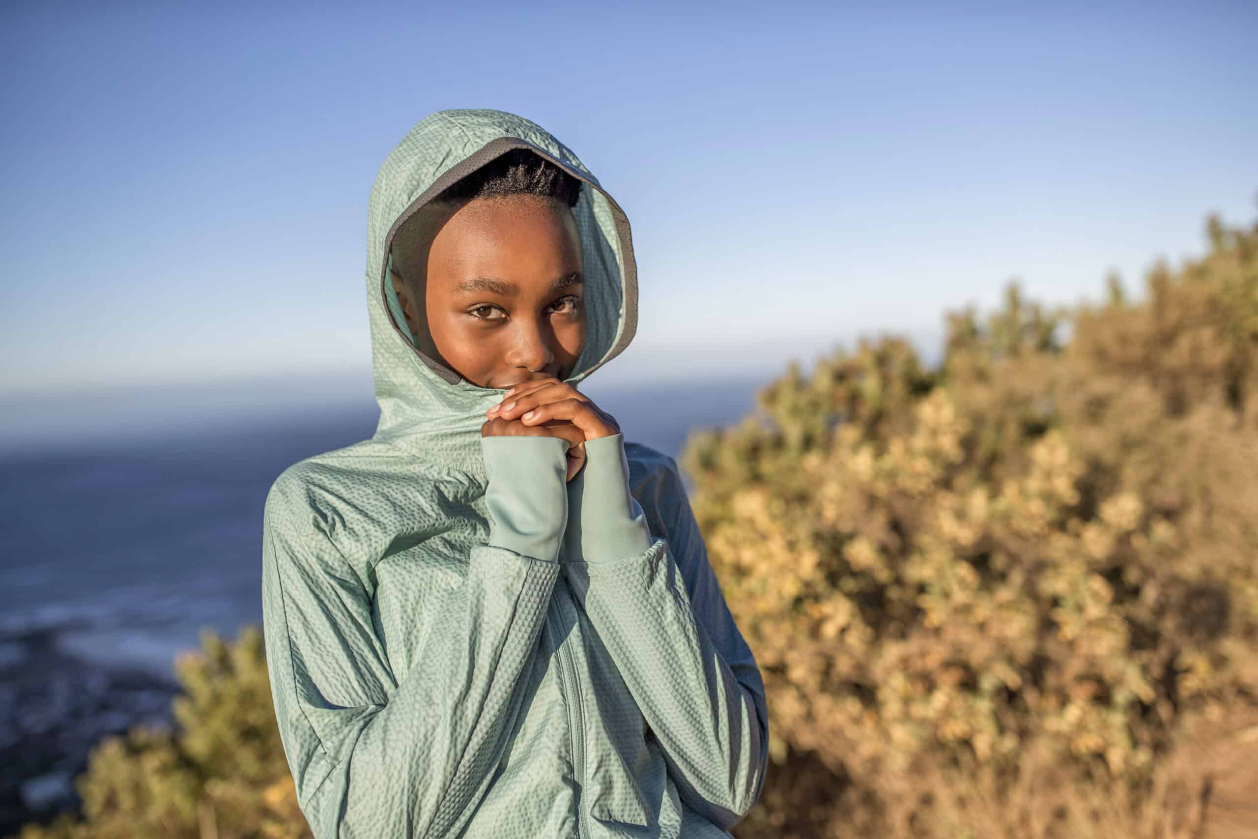 Eine junge Frau in einer hellblauen Kapuzenjacke steht im Freien, stützt ihr Kinn auf die Hand und lächelt sanft. Im Hintergrund ist sie von üppigem Grün und einem blauen Himmel umrahmt. © Fotografie Tomas Rodriguez