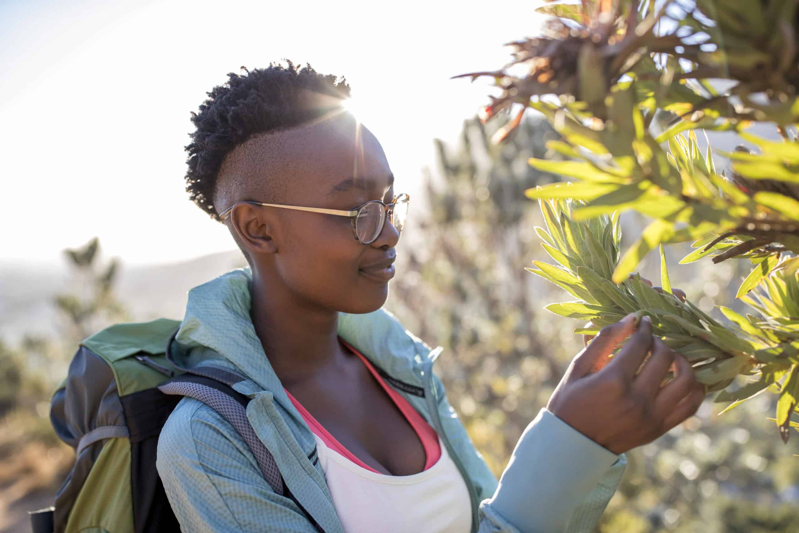Eine junge Frau mit kurzen Haaren, Brille, Rucksack und lässiger Outdoor-Kleidung untersucht während einer sonnigen Wanderung neugierig eine grüne Pflanze. © Fotografie Tomas Rodriguez