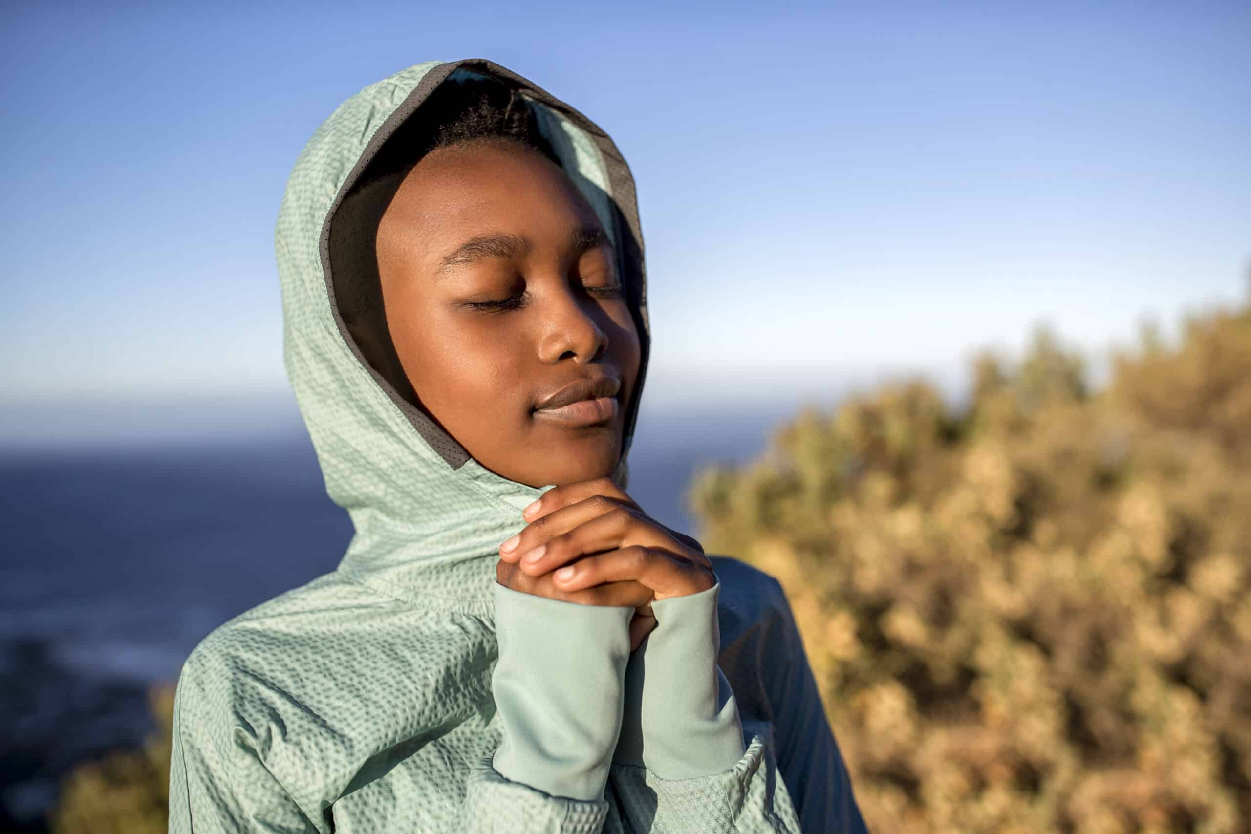 Eine gelassene junge Frau in einer blaugrünen Jacke mit Kapuze hält mit geschlossenen Augen sanft ihre Hände vor ihrem Gesicht zusammen und steht vor einem unscharfen natürlichen Hintergrund. © Fotografie Tomas Rodriguez