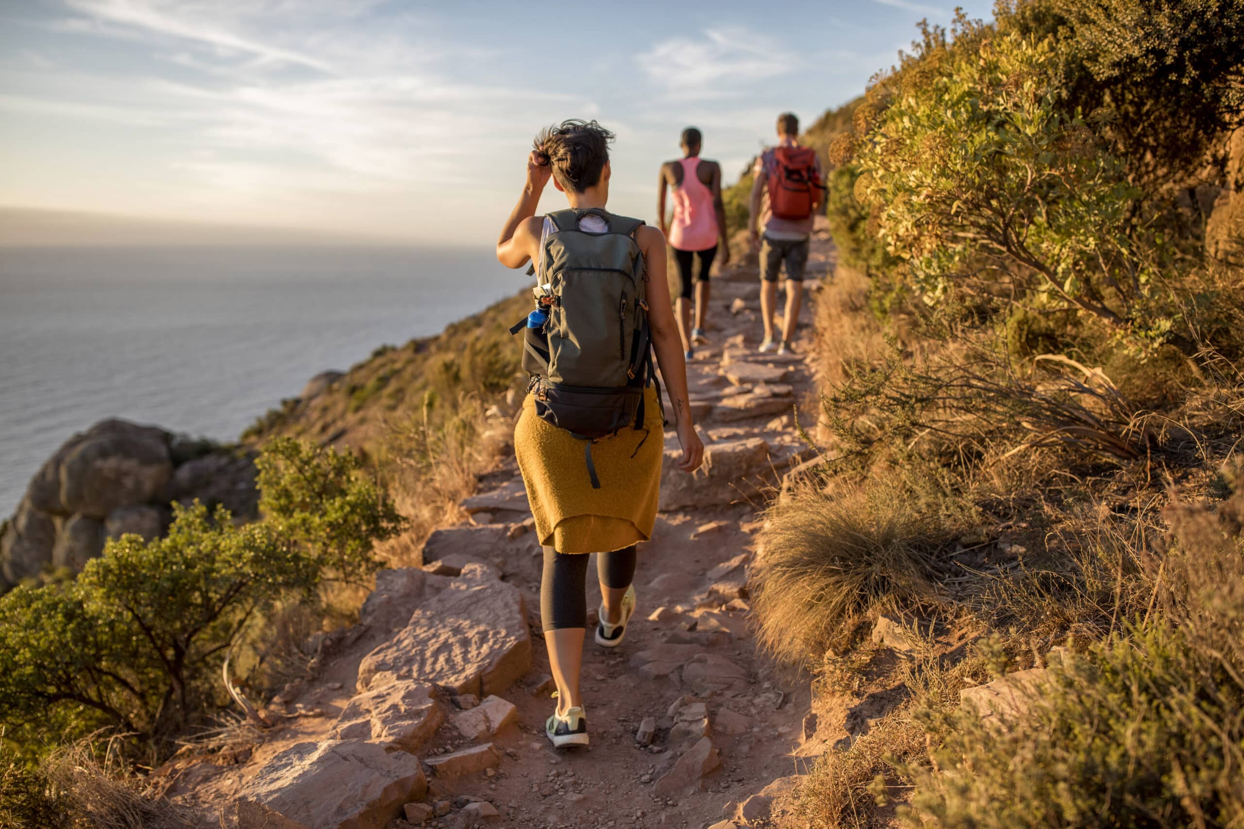 Drei Wanderer mit Rucksäcken wandern bei Sonnenuntergang auf einem felsigen Pfad, umgeben von üppigem Grün. Einer von ihnen trägt eine Beinprothese. © Fotografie Tomas Rodriguez