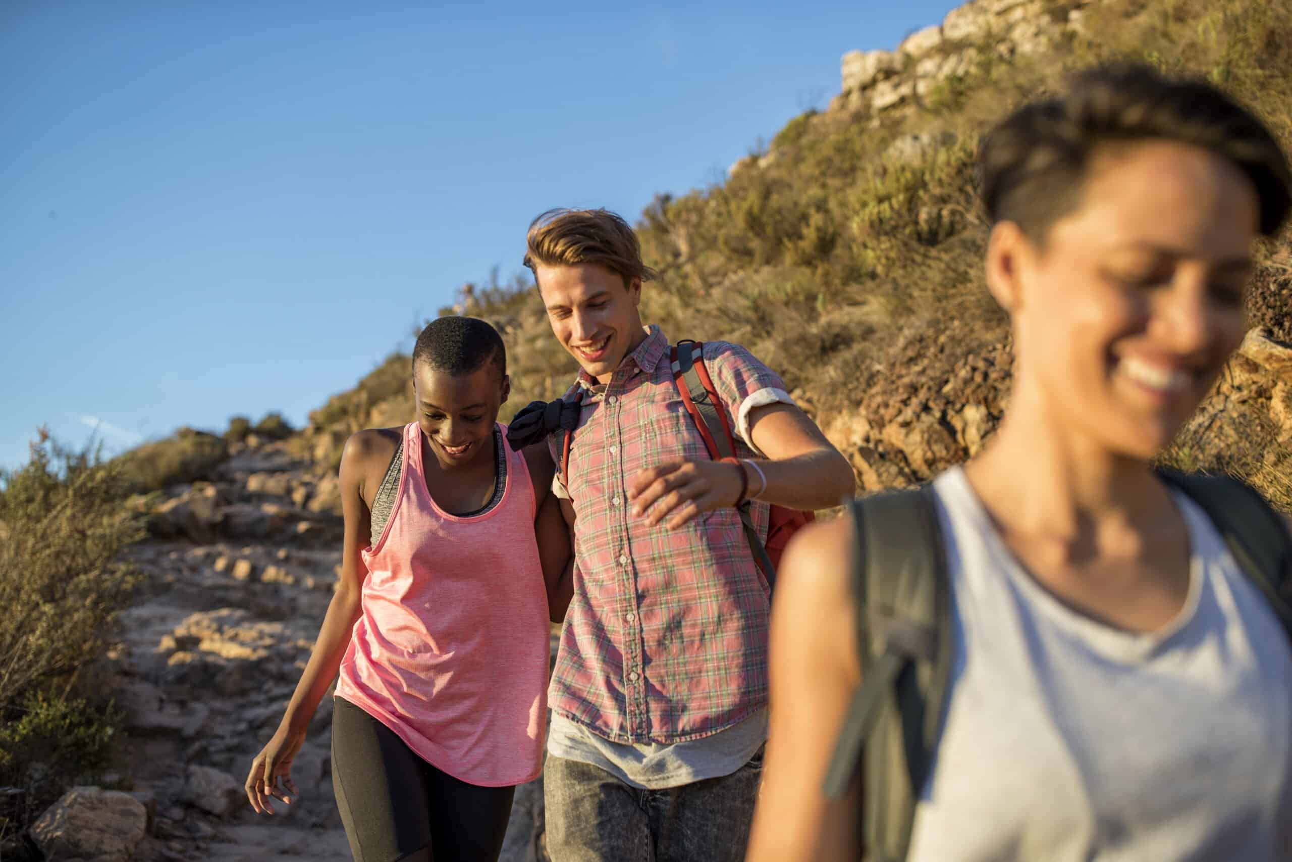 Drei Freunde wandern auf einem sonnigen Bergpfad, lächeln und plaudern, während sie gemeinsam durch eine felsige Landschaft gehen. Zwei Frauen und ein Mann genießen die Natur. © Fotografie Tomas Rodriguez