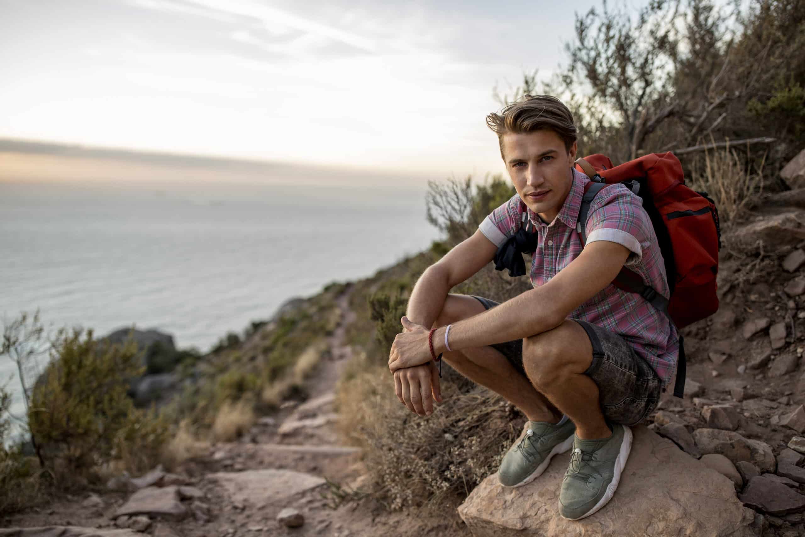 Ein junger Mann mit einem Rucksack sitzt auf einem Bergpfad und blickt in die Kamera. Im Hintergrund ist in der Abenddämmerung der ruhige Meereshorizont zu sehen. © Fotografie Tomas Rodriguez