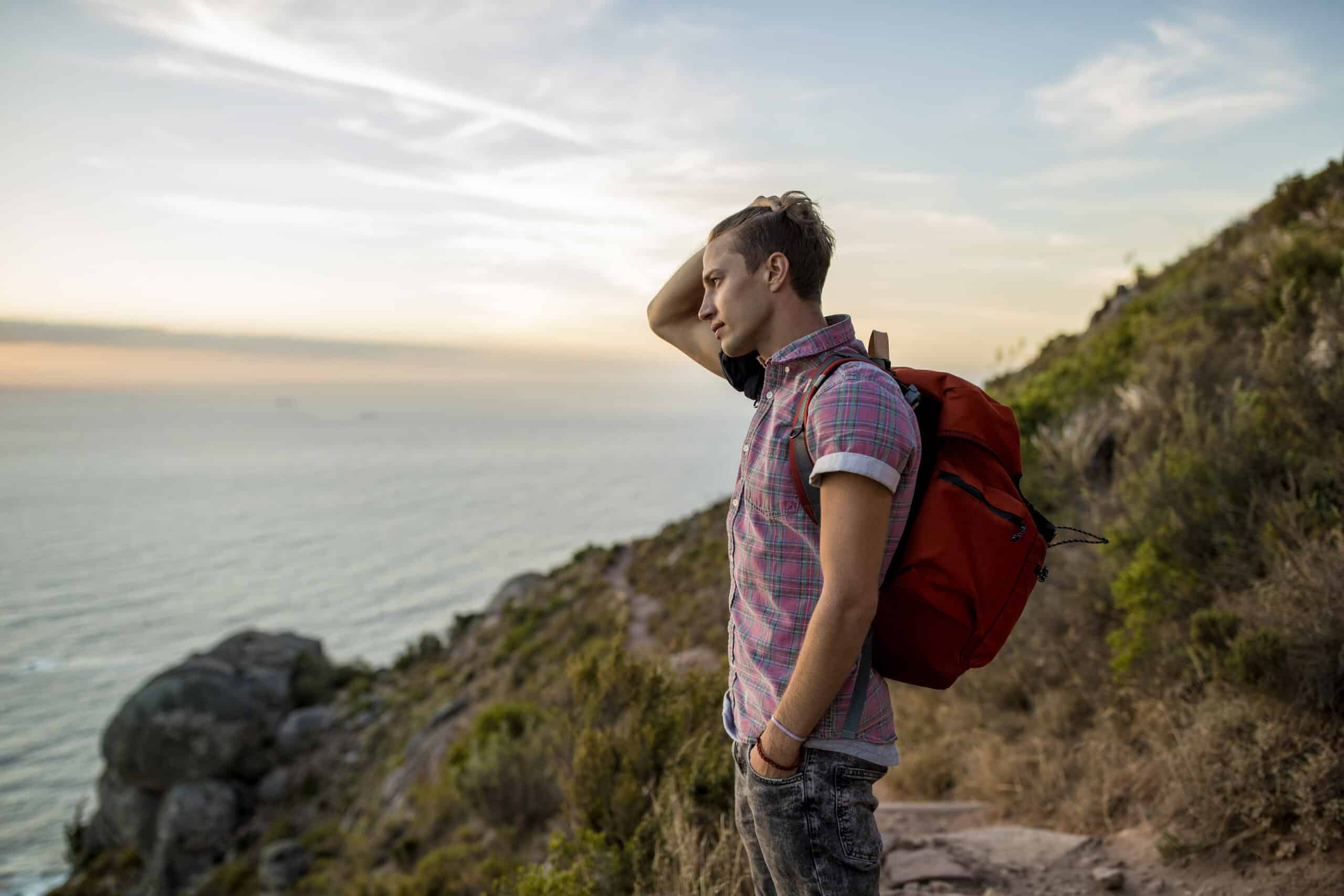 Ein junger Mann mit einem roten Rucksack steht auf einem Küstenpfad und blickt bei Sonnenuntergang auf das Meer, die Hand auf dem Kopf. © Fotografie Tomas Rodriguez