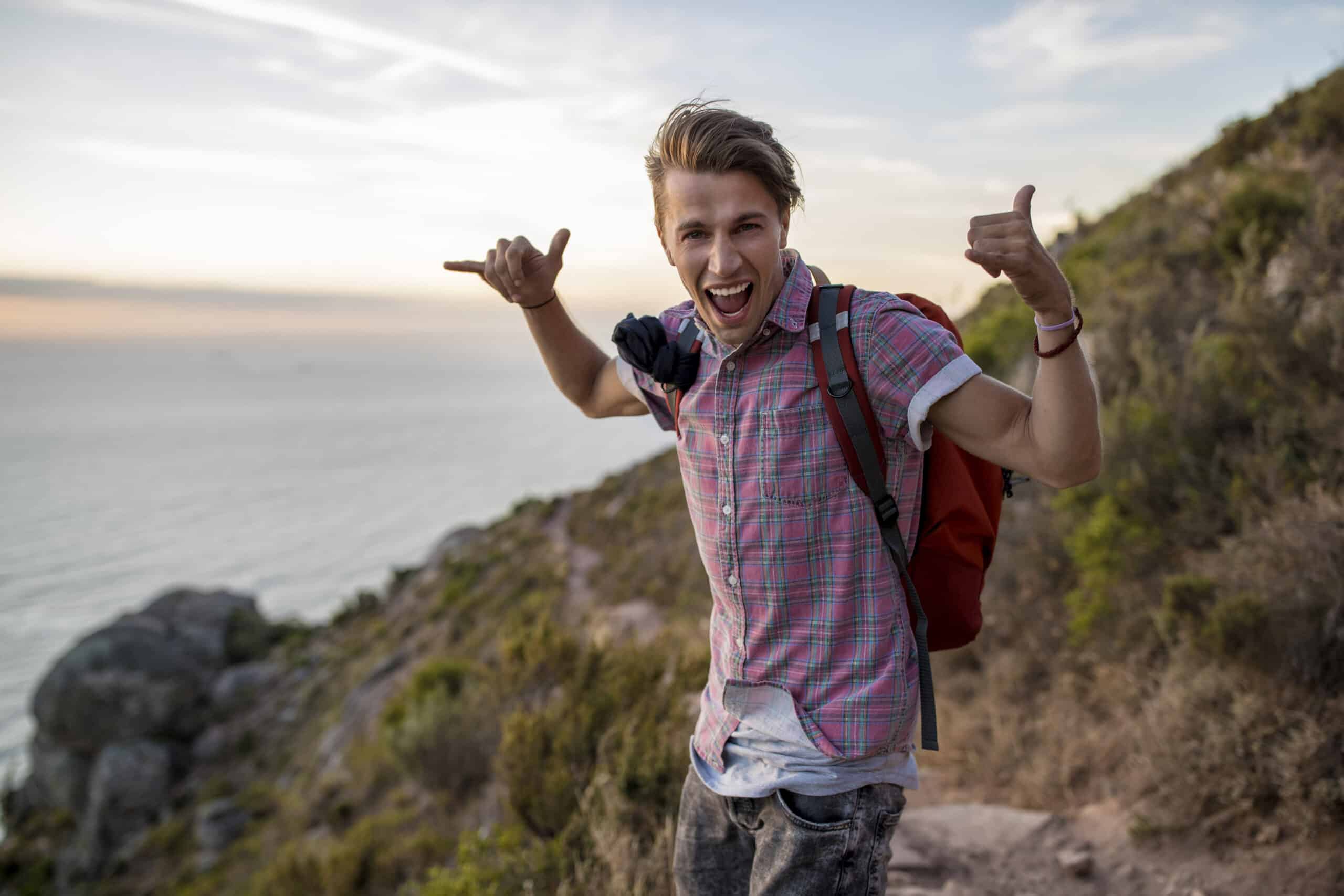 Ein fröhlicher junger Mann mit einem Rucksack streckt den Daumen nach oben und macht eine verspielte Geste. Er steht auf einem Küstenpfad mit dem Meer und den Klippen im Hintergrund bei Sonnenuntergang. © Fotografie Tomas Rodriguez