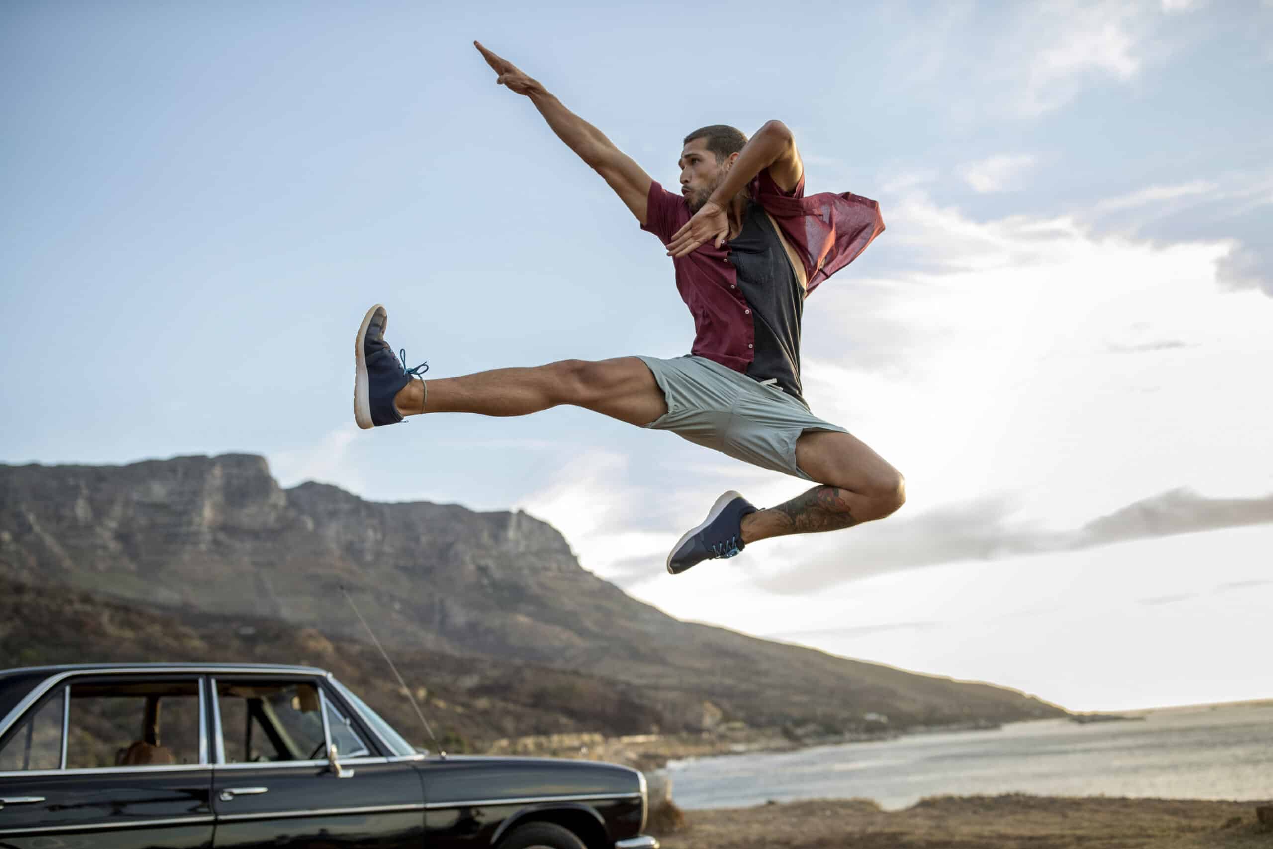Eine Person in der Luft, die mit ausgestreckten Armen einen dynamischen High Kick ausführt, vor einer malerischen Kulisse aus Bergen und einer Küste. Links parkt ein Oldtimer. Der Himmel ist teilweise bewölkt, die Sonne wirft einen warmen Schein. © Fotografie Tomas Rodriguez
