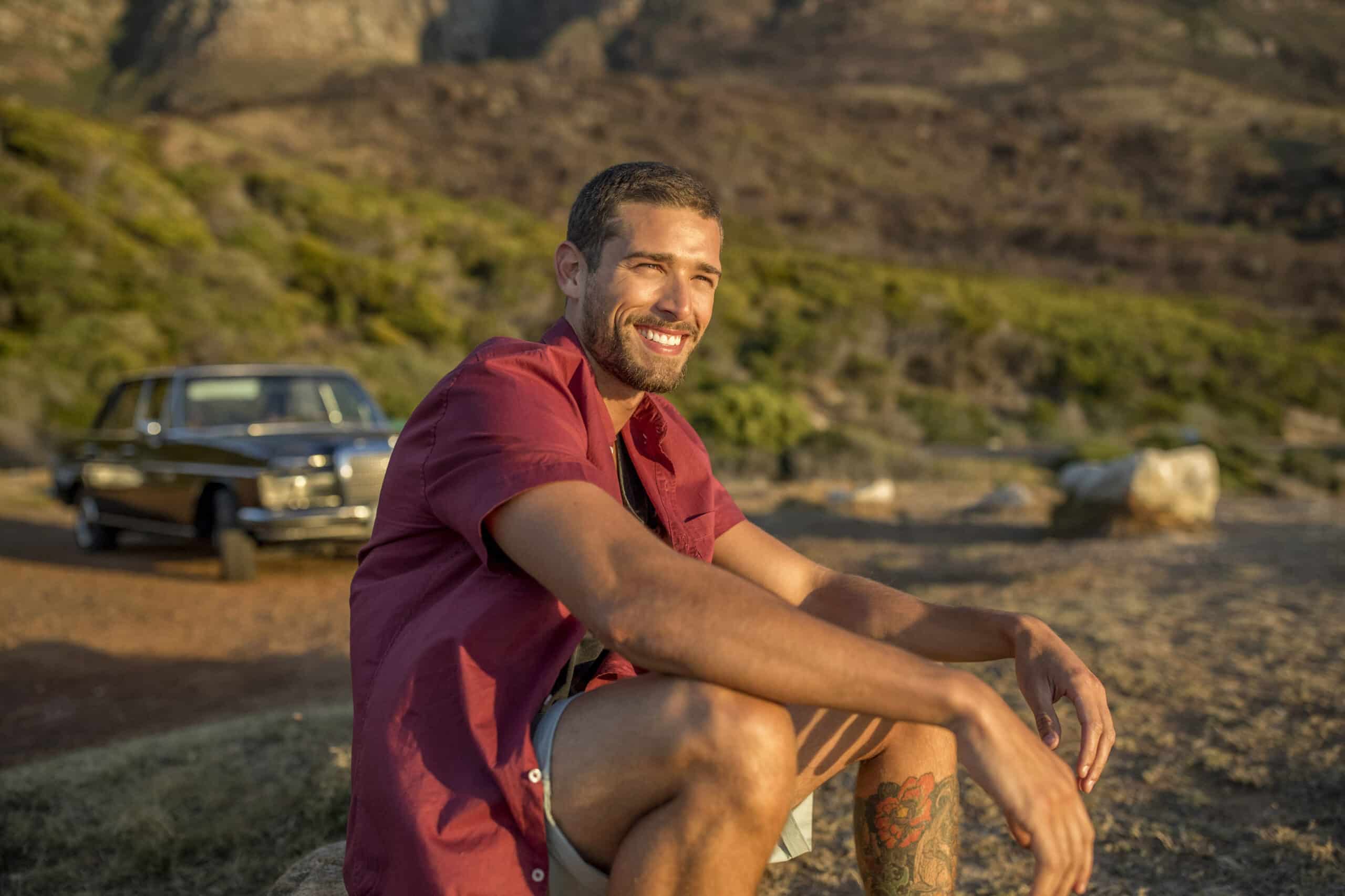 Ein Mann mit Bart und Tattoos auf dem linken Bein sitzt lächelnd auf einem Felsen im Freien. Er trägt ein rotes Hemd und Shorts. Im Hintergrund sind ein altes Auto und malerische Hügel zu sehen, die in warmes Sonnenlicht getaucht sind. © Fotografie Tomas Rodriguez