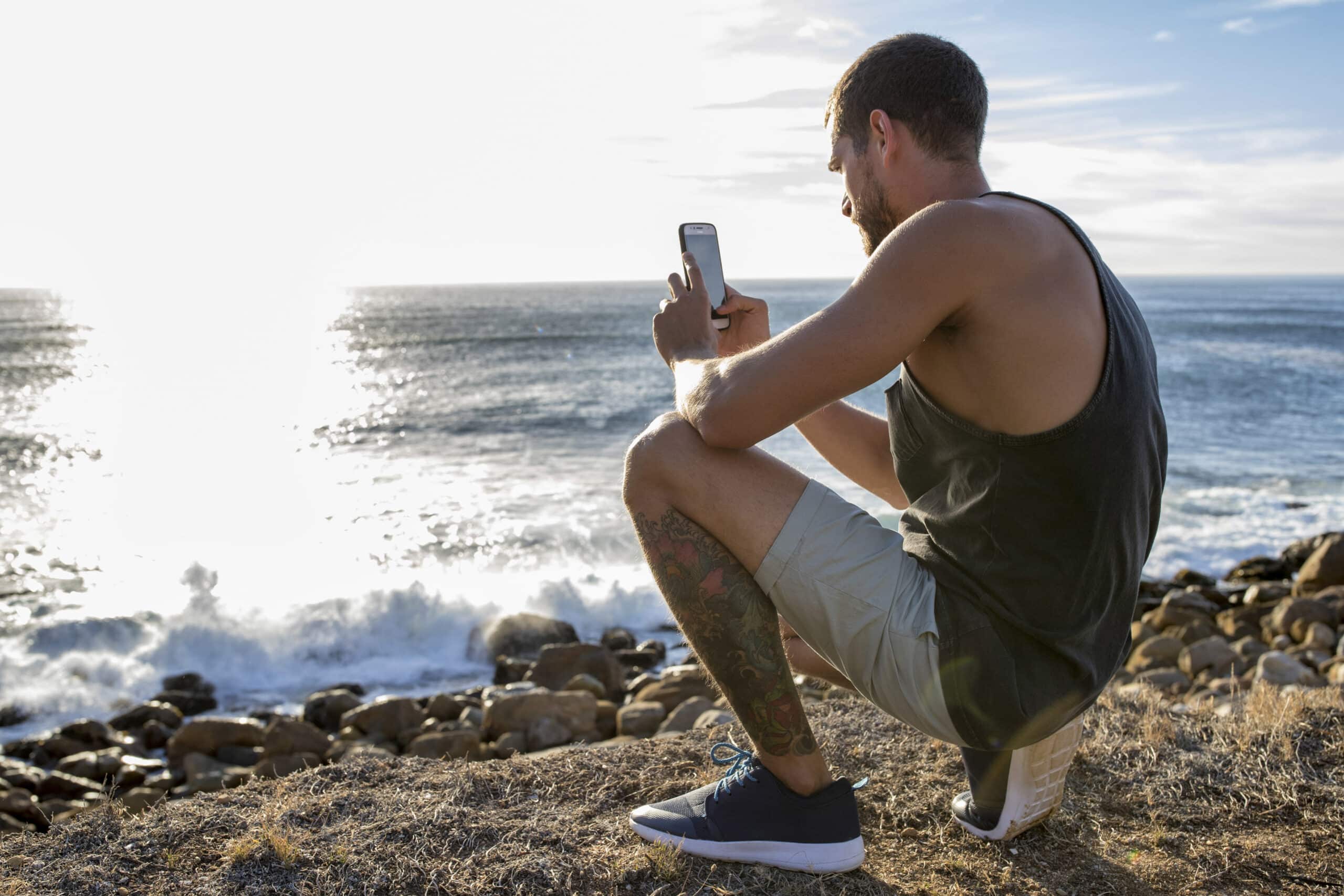 Ein Mann mit tätowiertem Bein, der ein Tanktop und Shorts trägt, hockt auf einer felsigen Strandklippe und macht mit seinem Telefon ein Foto vom Meer. Die Sonne scheint hell über das Wasser und schafft eine ruhige und malerische Szene. © Fotografie Tomas Rodriguez