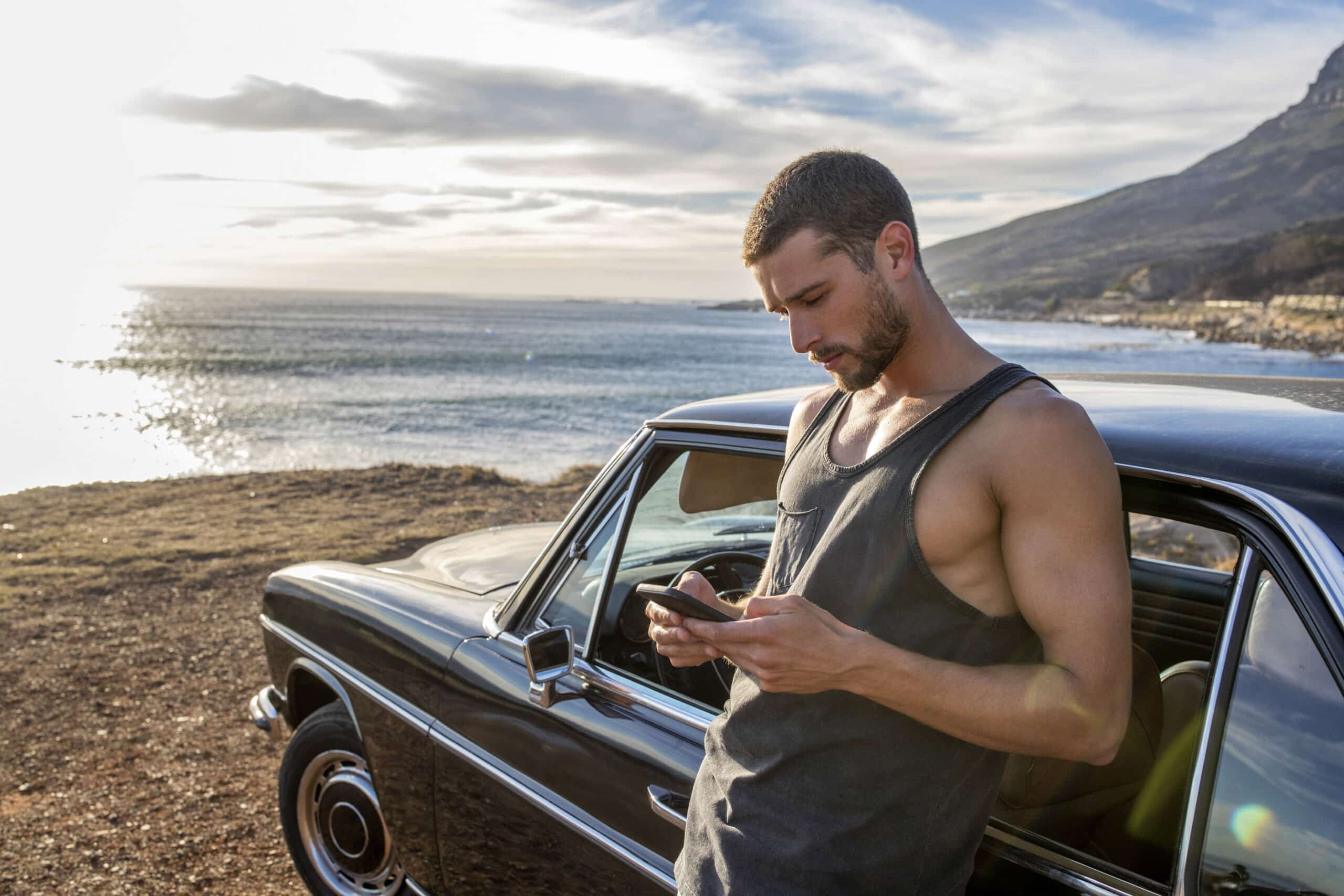 Ein Mann in einem ärmellosen Hemd steht neben einem geparkten schwarzen Auto und schaut auf sein Telefon. Der Standort scheint an der Küste zu sein, mit dem Meer und einem klaren Himmel im Hintergrund. © Fotografie Tomas Rodriguez