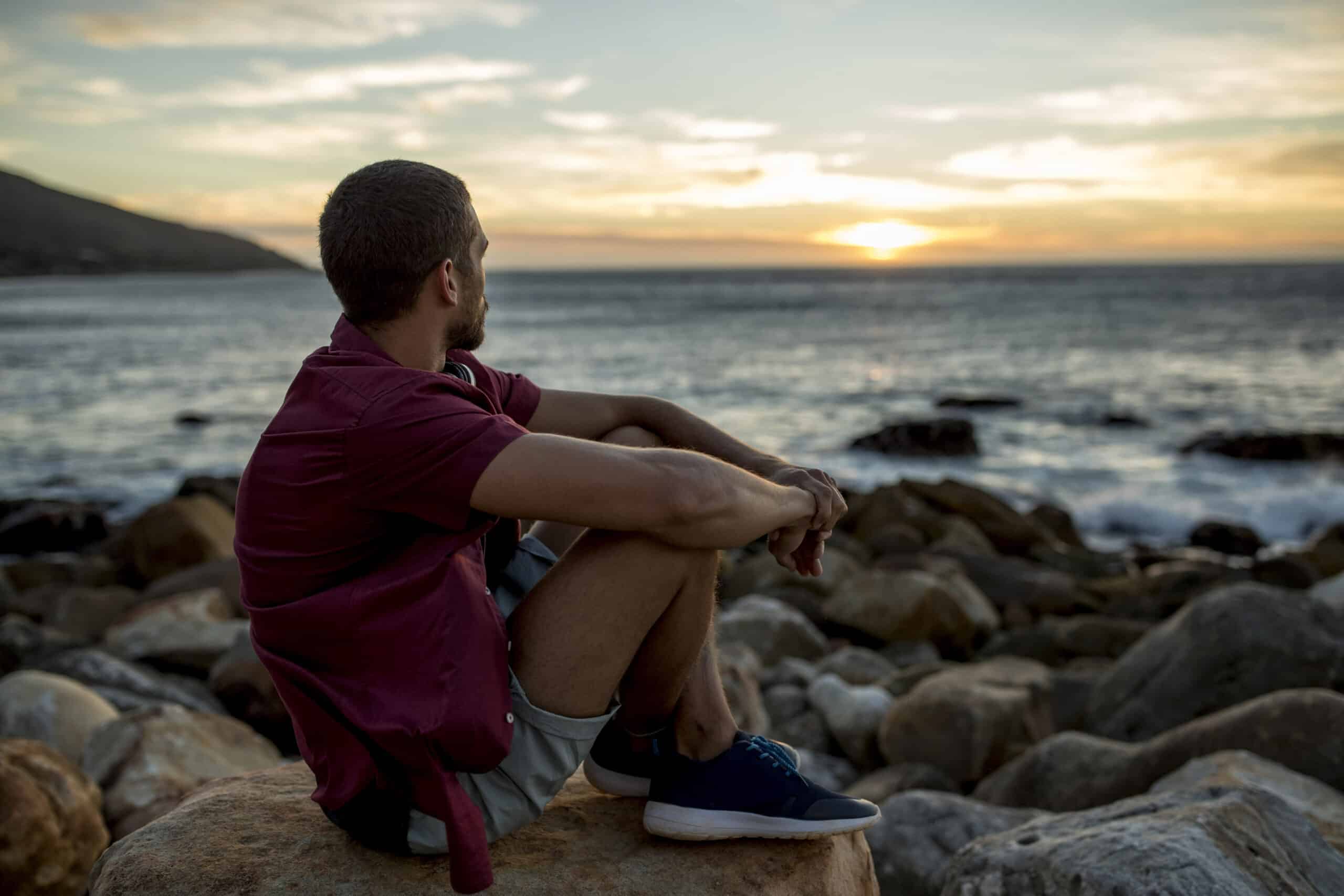 Ein Mann mit kurzen Haaren und Bart sitzt auf einem Felsen am Strand und blickt bei Sonnenuntergang auf das Meer. Er trägt ein kastanienbraunes Hemd, graue Shorts und blaue Turnschuhe. Der Himmel ist in warmen Farbtönen gefärbt, während die Sonne hinter dem Horizont untergeht. © Fotografie Tomas Rodriguez