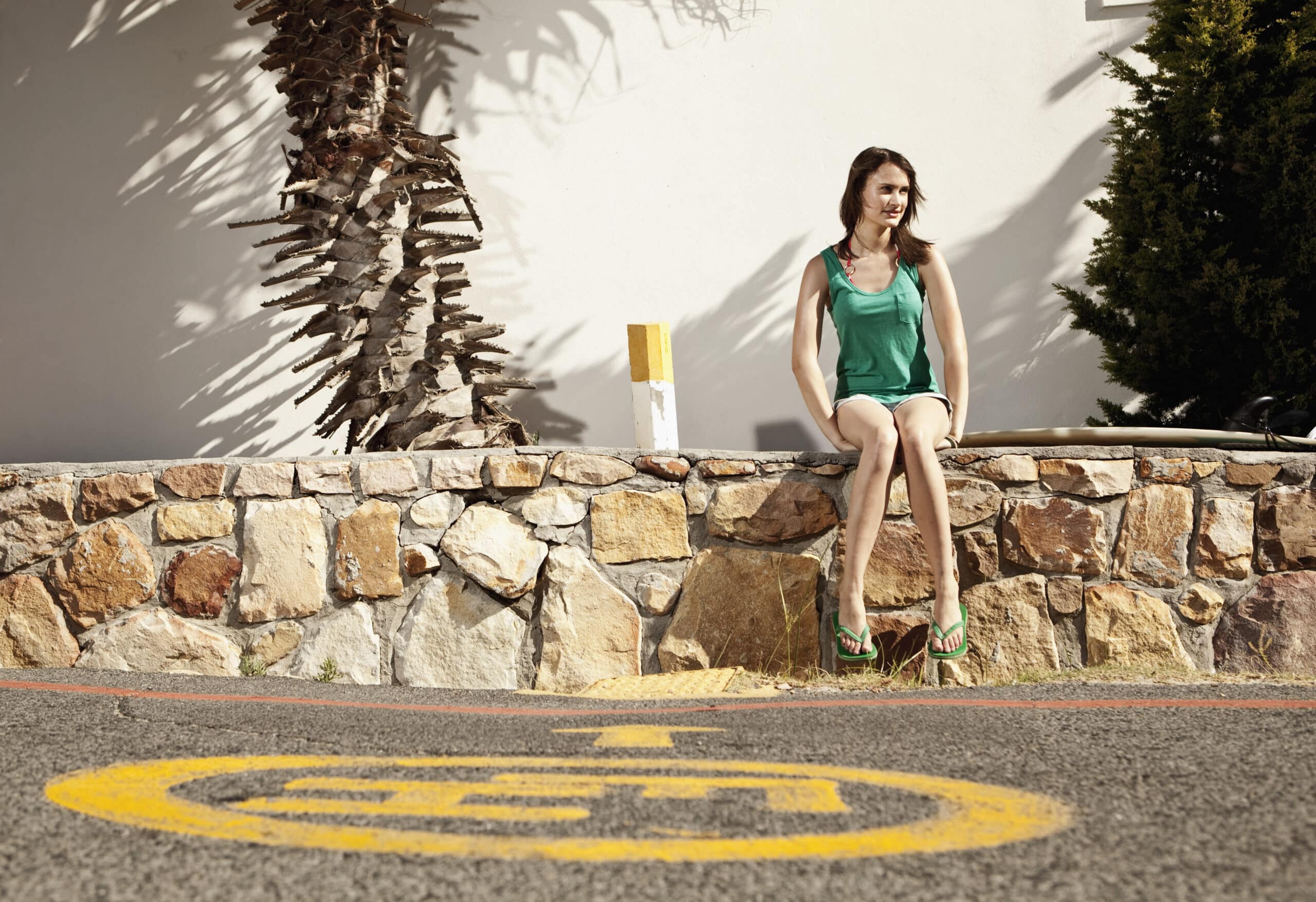 Eine Frau in einem grünen Kleid und grünen Flip-Flops sitzt auf einer Steinmauer in der Nähe einer Straße mit einem gelben runden Symbol und Buchstaben. Im Hintergrund sind eine große, vertrocknete Palme und ein grüner Busch zu sehen. Die Szene ist sonnig und wirft Schatten auf die Mauer und den Boden. © Fotografie Tomas Rodriguez