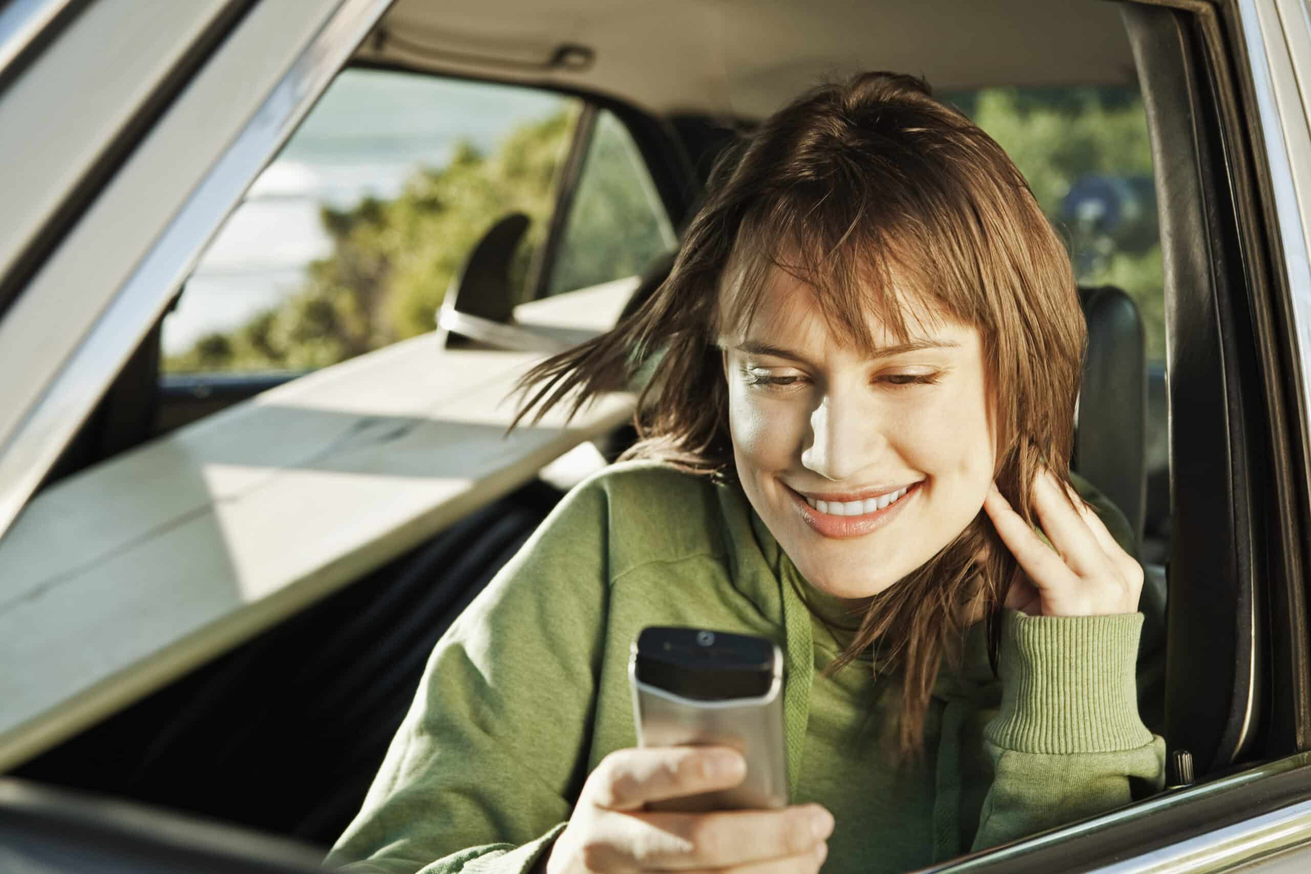 Eine Person mit schulterlangem Haar sitzt in einem Auto und lächelt, während sie auf ein Smartphone schaut. Sie trägt einen grünen Kapuzenpullover und hat ihre Hand neben dem Kopf. Auf dem Rücksitz des Autos ist ein Surfbrett zu sehen. Im Hintergrund ist üppiges Grün zu sehen. © Fotografie Tomas Rodriguez