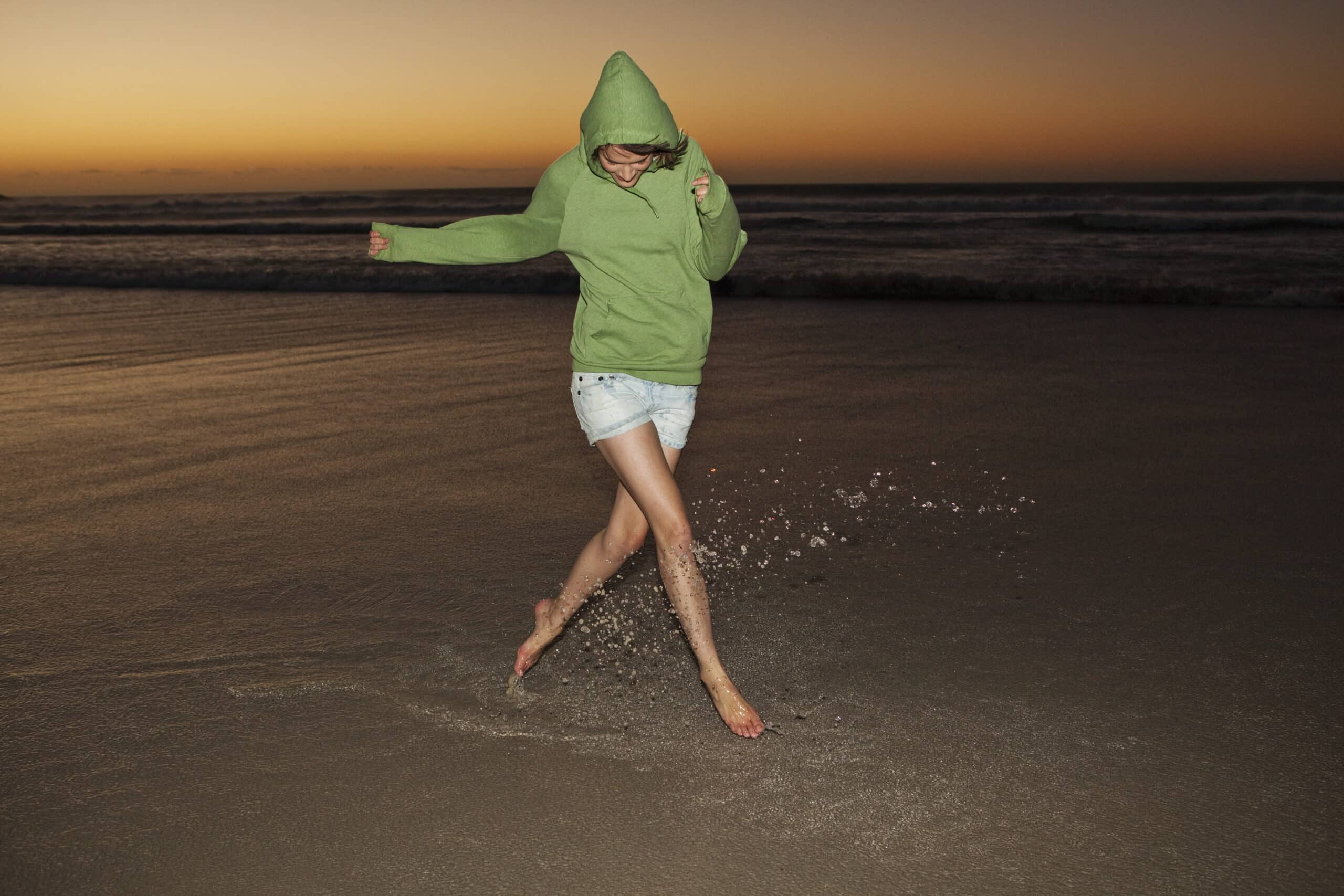 Eine Person mit einem grünen Kapuzenpullover und Jeansshorts planscht fröhlich im Wasser an einem Strand bei Sonnenuntergang. Der Himmel ist in warmen Orange- und Rosatönen gehalten, während die Wellen eine ruhige Kulisse bilden. © Fotografie Tomas Rodriguez