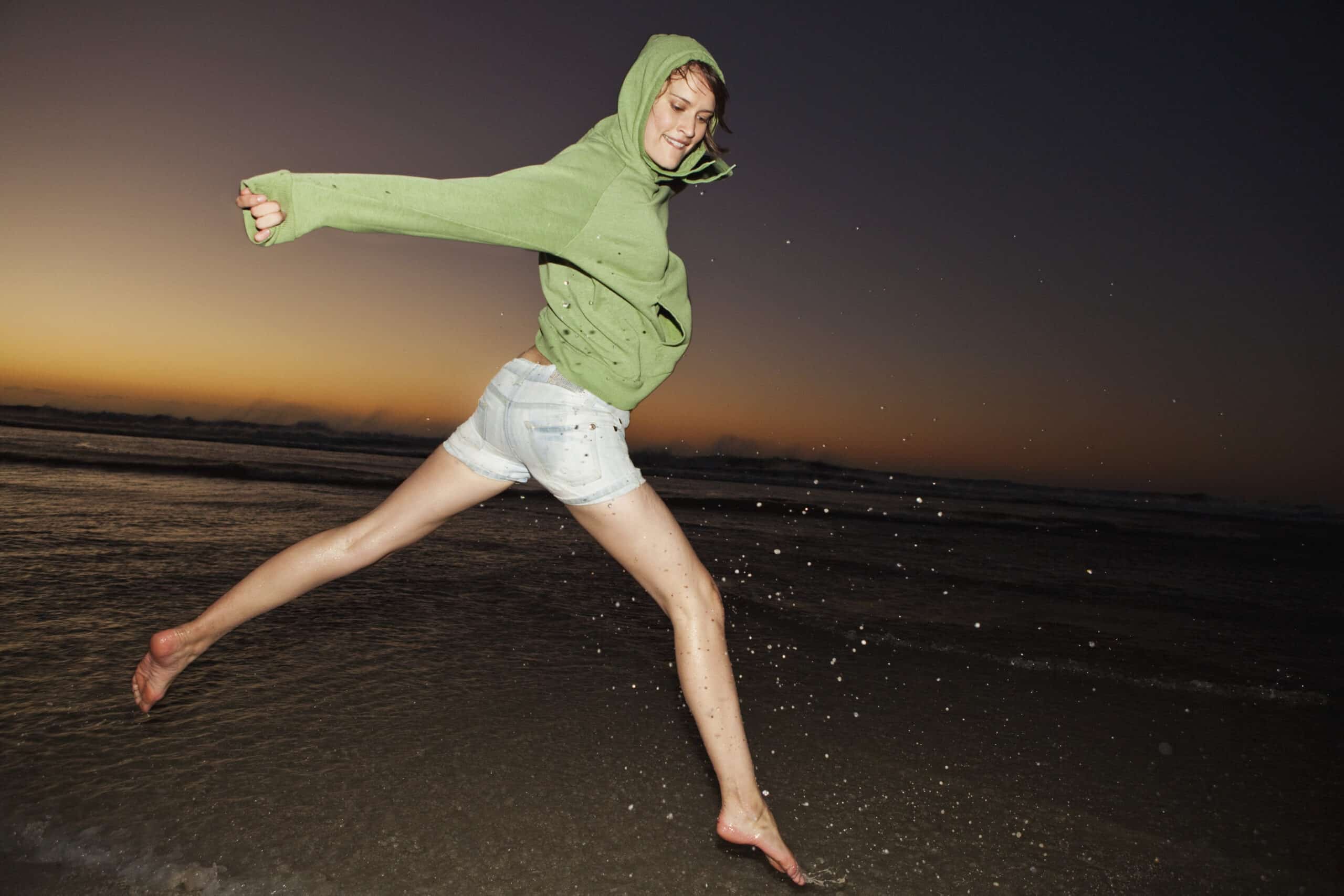 Eine Person in einem grünen Kapuzenpullover und Jeansshorts springt in der Abenddämmerung fröhlich am Strand herum. Der Himmel geht vom Sonnenuntergang in leuchtende Farben über, die am Horizont verschwinden. Spritzendes Wasser spritzt hinter ihren Füßen her, während sie den Moment genießt. © Fotografie Tomas Rodriguez