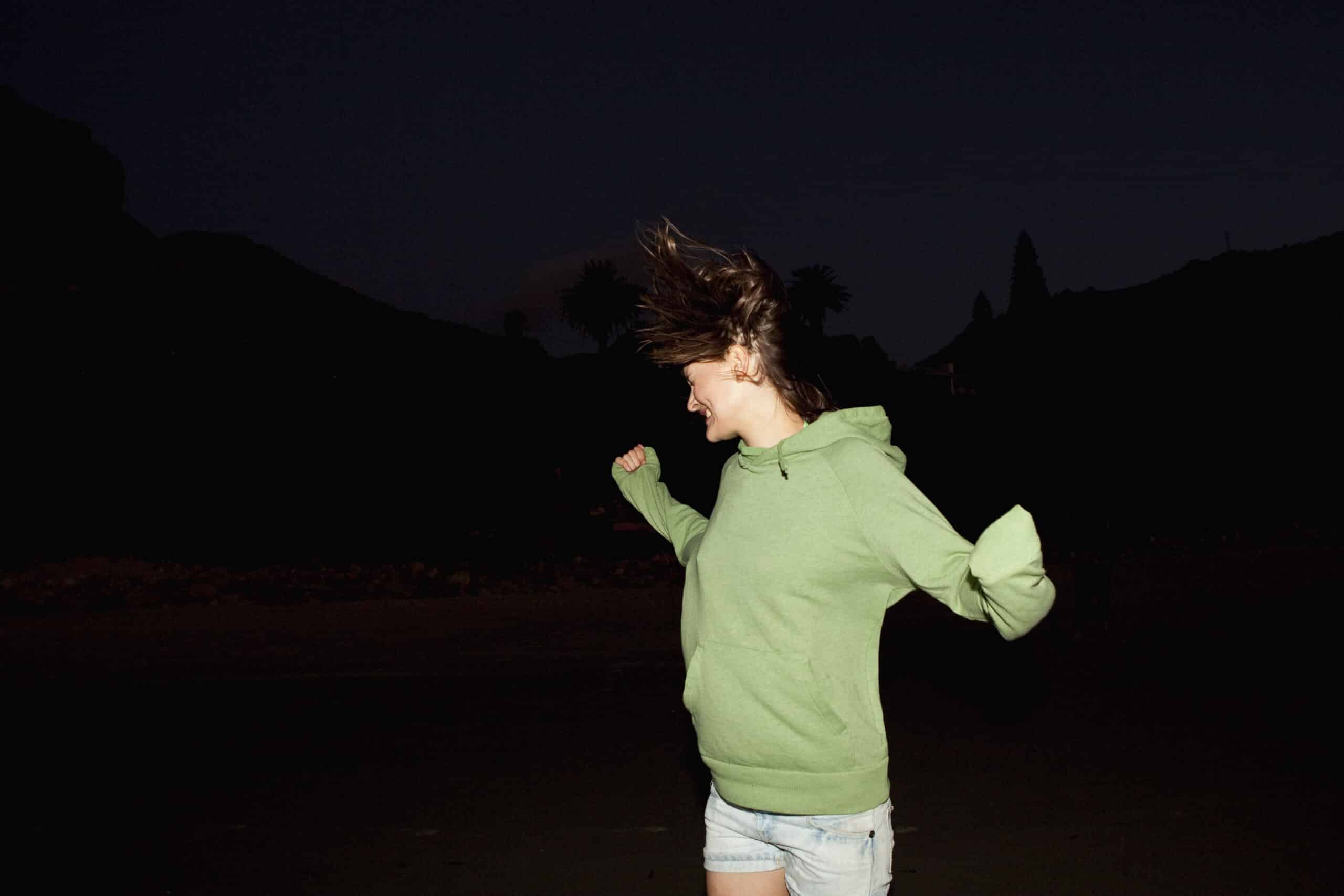 Frau mit grünem Kapuzenpullover und Jeansshorts steht nachts im Freien. Ihr Haar weht zur Seite und sie scheint in Bewegung zu sein. Der Hintergrund ist dunkel mit schwachen Silhouetten von Bäumen und Bergen. © Fotografie Tomas Rodriguez