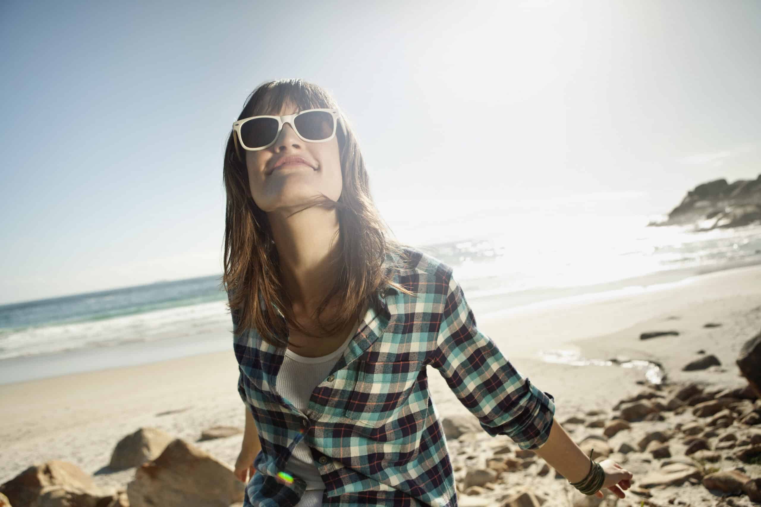 Eine Frau mit weißer Sonnenbrille und kariertem Hemd steht lächelnd an einem steinigen Strand. Die Sonne scheint hell am klaren blauen Himmel und im Hintergrund plätschern die Meereswellen sanft ans Ufer. © Fotografie Tomas Rodriguez