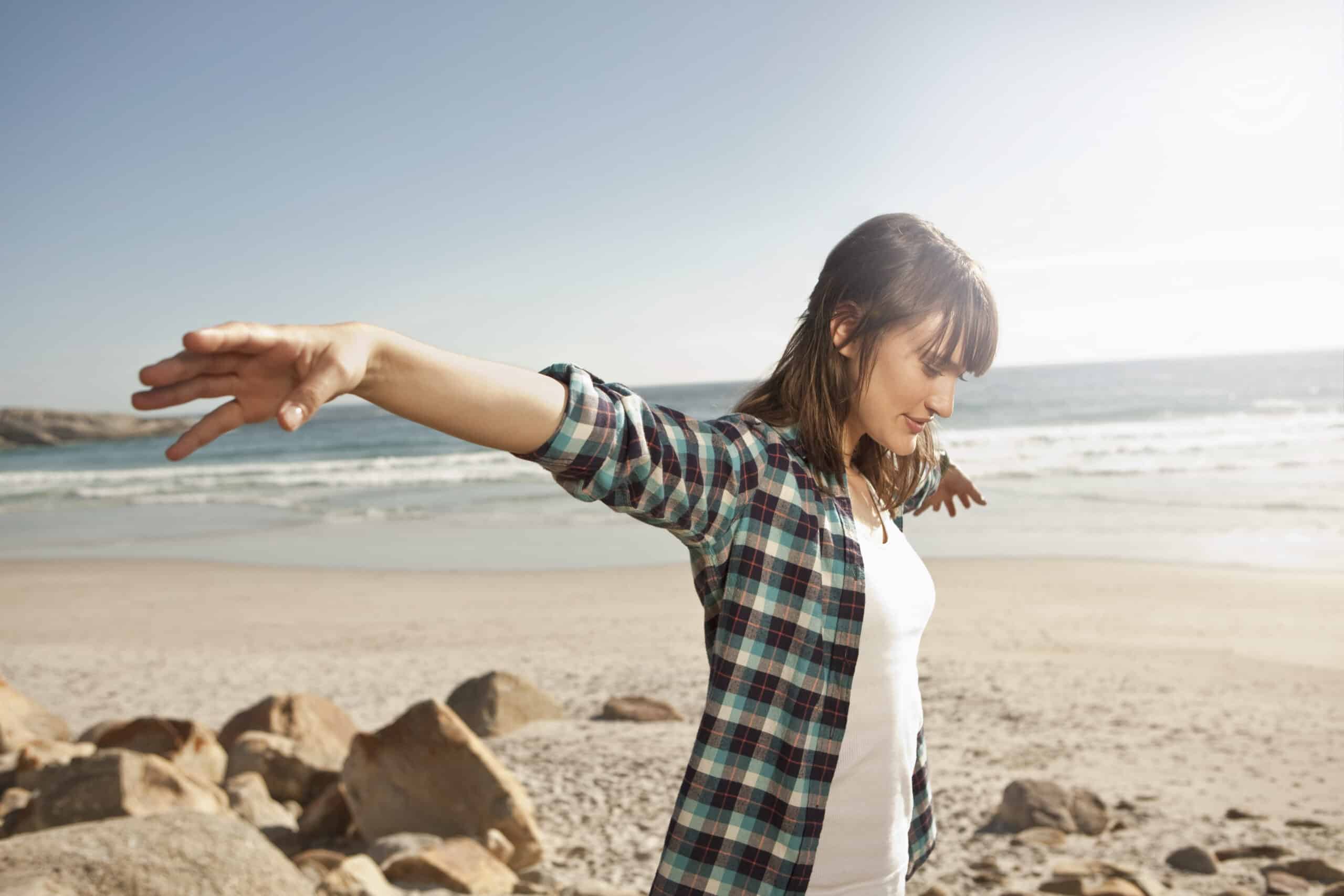 Eine Person mit schulterlangem Haar in einem karierten Hemd und einem weißen T-Shirt steht mit ausgestreckten Armen an einem Strand. Sie scheint das sonnige Wetter und die Meereswellen im Hintergrund zu genießen. Im Vordergrund sind Felsen zu sehen. © Fotografie Tomas Rodriguez