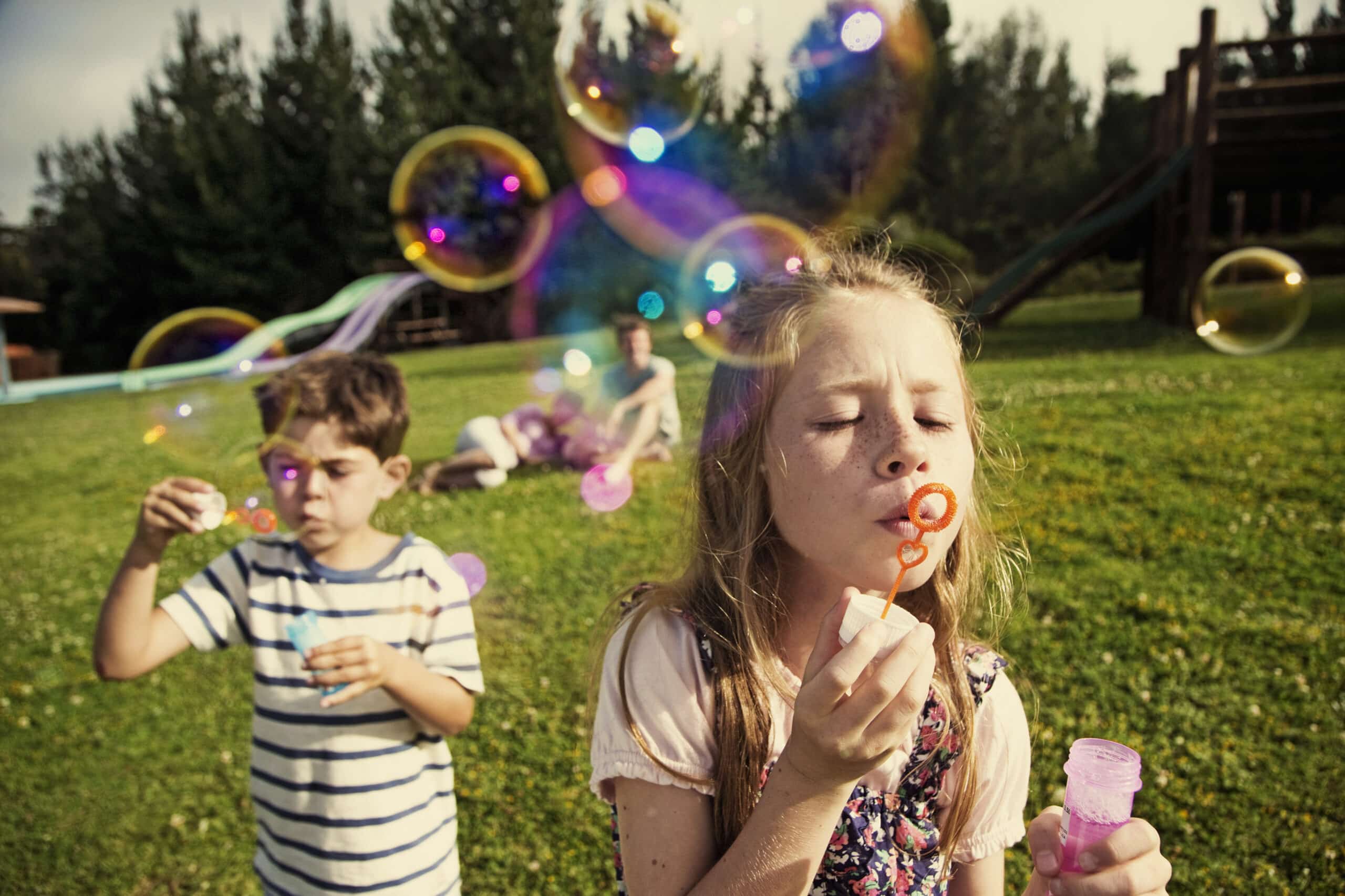 Ein junges Mädchen und ein Junge pusten Seifenblasen in einem sonnigen Park. Im Hintergrund schweben bunte Seifenblasen durch die Luft und im Hintergrund spielen Kinder auf einem Spielplatz. © Fotografie Tomas Rodriguez