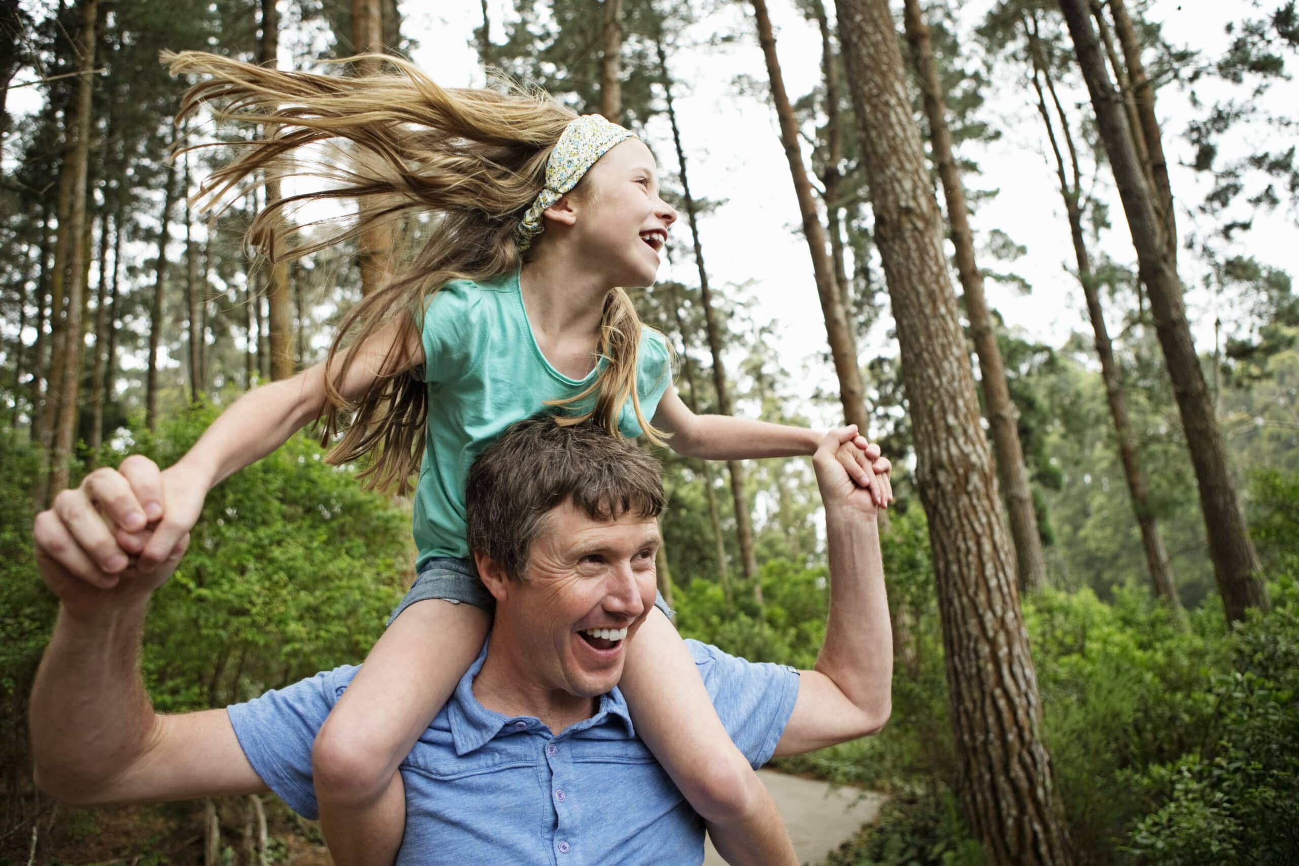 Ein fröhliches junges Mädchen mit wehendem Haar reitet auf den Schultern eines Mannes, beide lachen in einer üppigen Waldlandschaft. © Fotografie Tomas Rodriguez