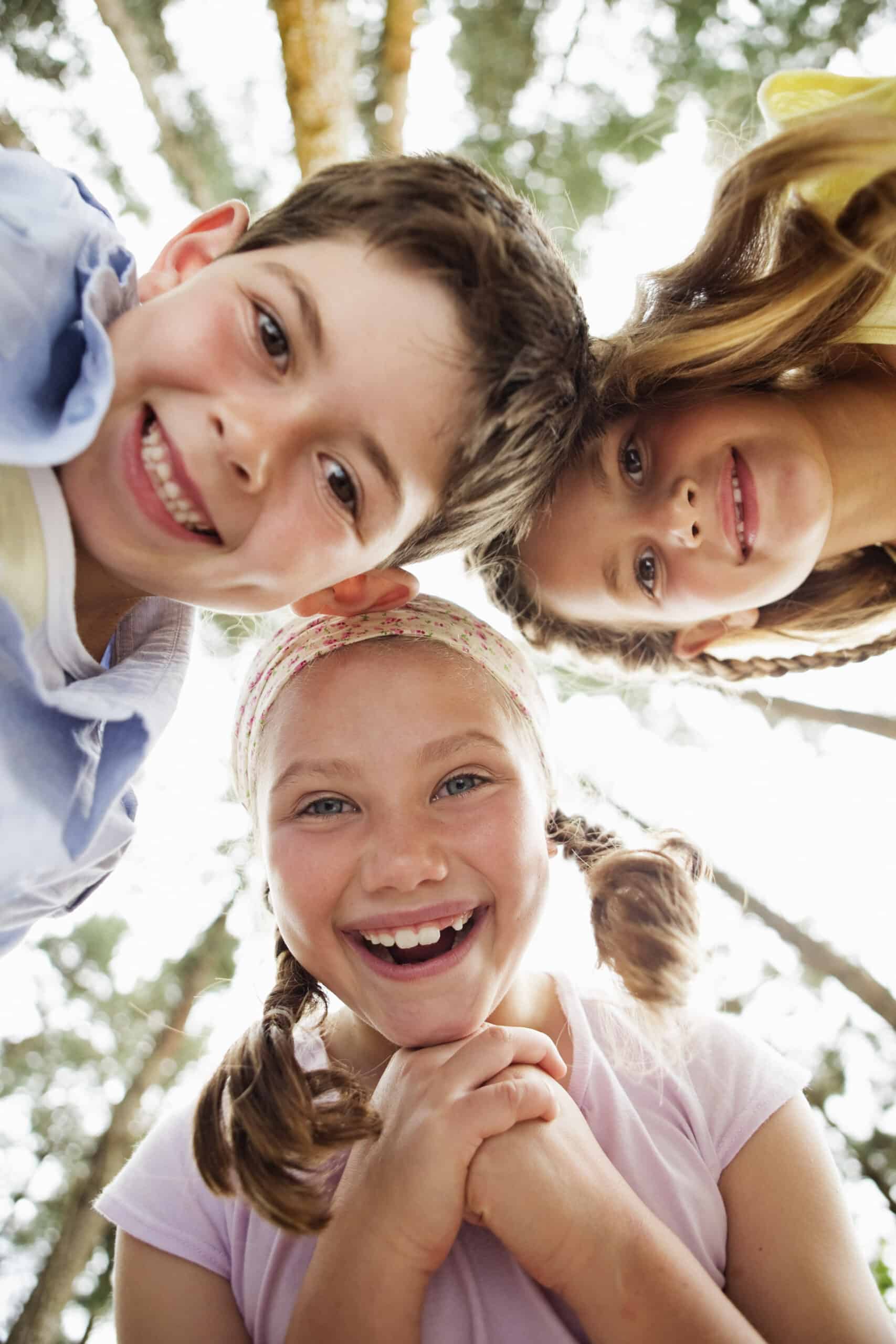 Drei lächelnde Kinder blicken von oben auf die Kamera, im Hintergrund sind die Bäume unscharf, und fangen einen freudigen, unbeschwerten Moment unter Freunden im Freien ein. © Fotografie Tomas Rodriguez
