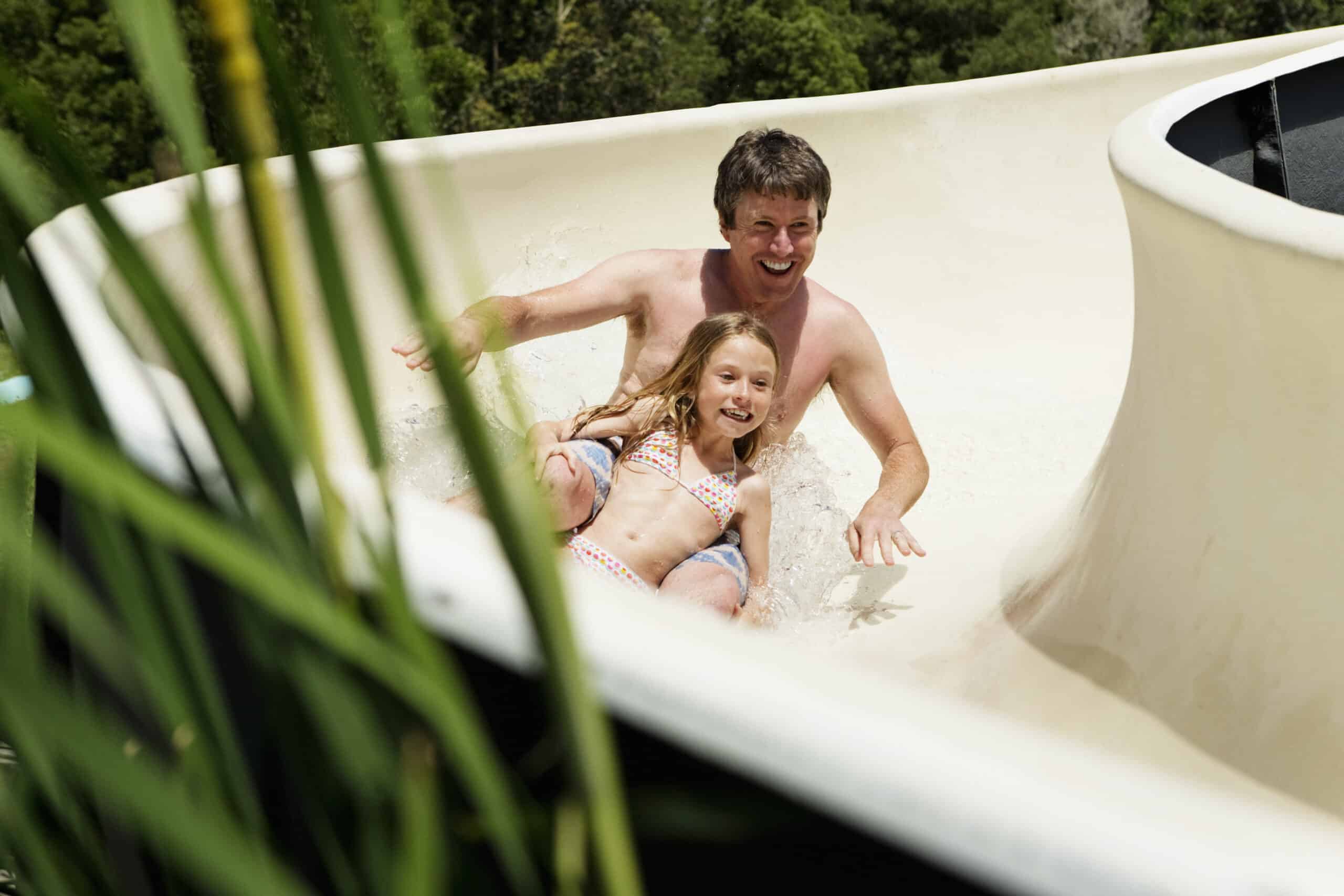 Ein fröhlicher Mann und ein junges Mädchen rutschen umgeben von üppigem Grün eine Wasserrutsche hinunter und lachen beide vor Aufregung. © Fotografie Tomas Rodriguez