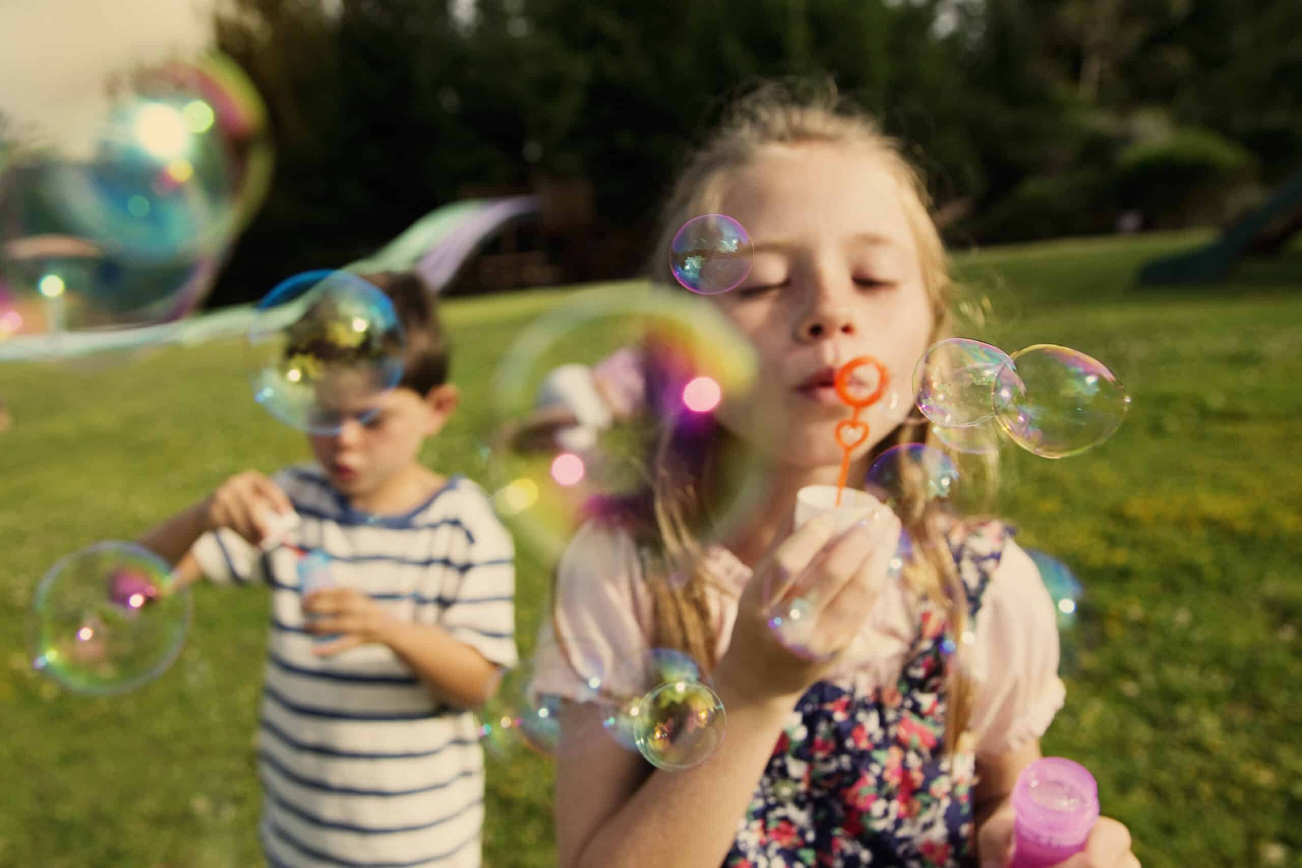 Im Fokus ist ein junges Mädchen zu sehen, das mit einem Stab Seifenblasen bläst, während im Hintergrund ein Junge ebenfalls mit Blasen spielt; das Ganze in einem sonnigen Park mit grünem Gras. © Fotografie Tomas Rodriguez