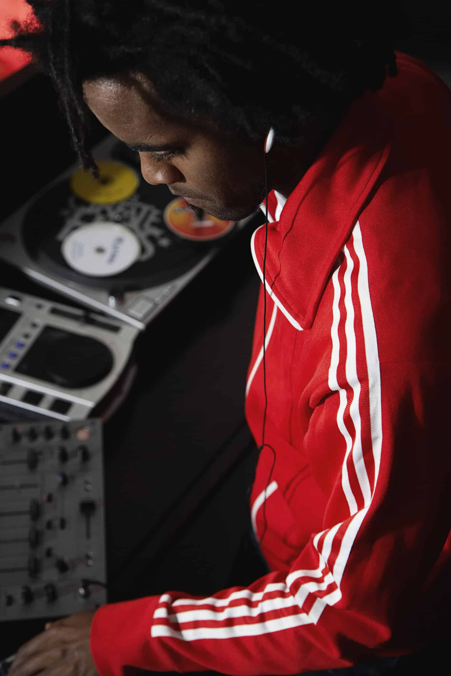 Ein DJ trägt einen roten Trainingsanzug mit weißen Streifen und Kopfhörer und mixt Musik auf professionellem DJ-Equipment. Er ist konzentriert dabei, die Regler einzustellen. Die Beleuchtung ist gedämpft, sodass das Equipment und seine Kleidung gut zur Geltung kommen. © Fotografie Tomas Rodriguez