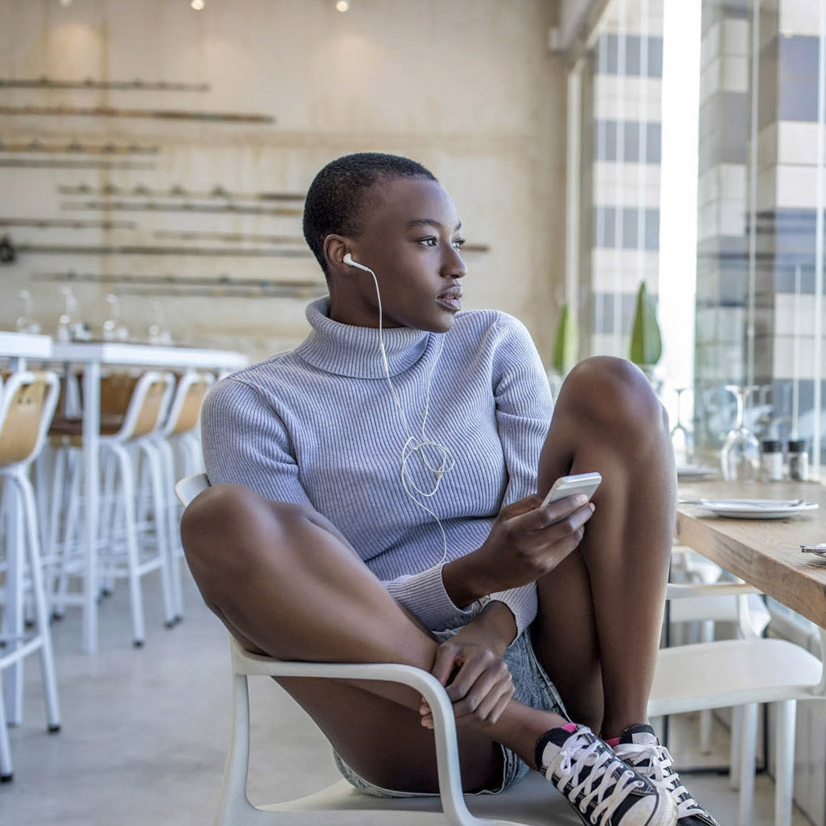 Eine Person mit Kurzhaarschnitt, grauem Rollkragenpullover und Jeansshorts sitzt auf einem Stuhl in einem modernen Café. Sie trägt Ohrstöpsel, hält ein Smartphone in der Hand und blickt nachdenklich aus dem Fenster. Das Café hat weiße Barhocker und ist minimalistisch eingerichtet. © Fotografie Tomas Rodriguez