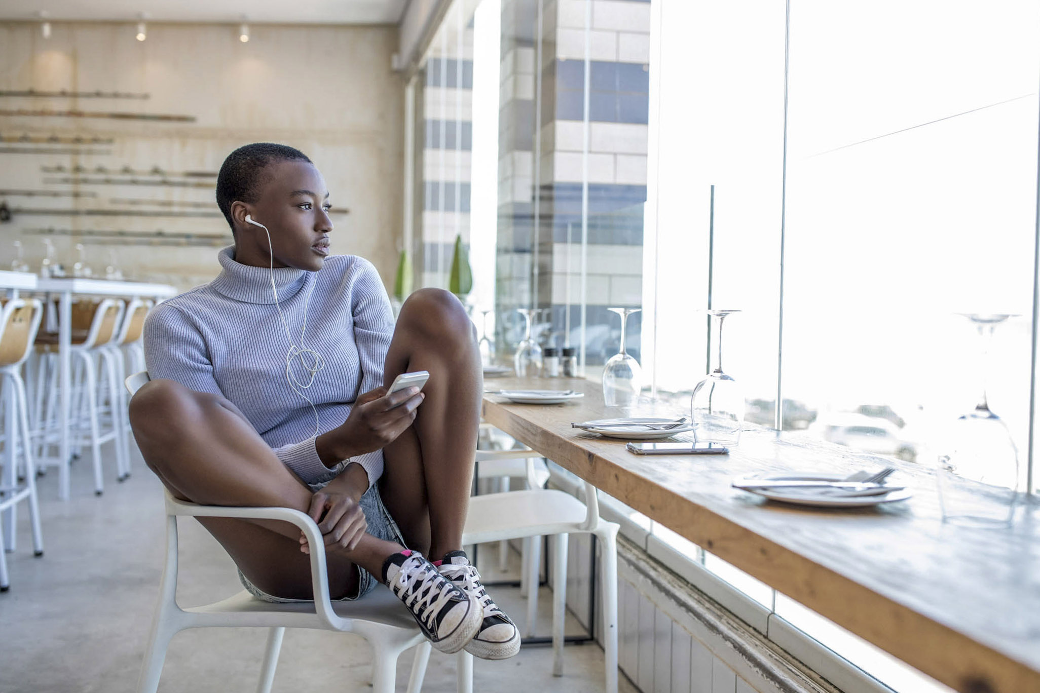 Eine Person mit kurzen Haaren, einem leichten Pullover und Shorts sitzt in einem Restaurant und hat die Beine auf einem Stuhl hochgelegt. Sie hört über Kopfhörer etwas und blickt nachdenklich aus einem großen Fenster. Im Hintergrund sind leere Tische mit Gedecken zu sehen. © Fotografie Tomas Rodriguez