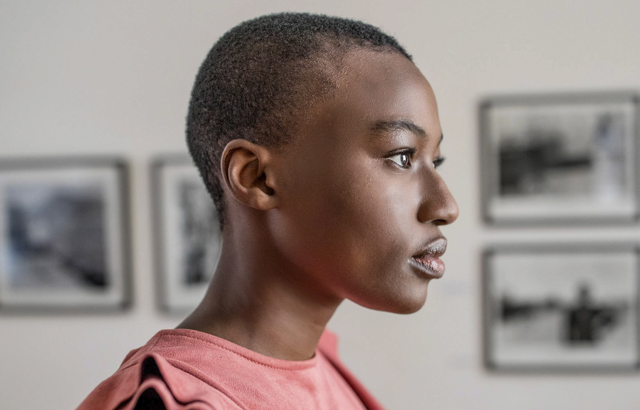 Eine Person mit kurzen Haaren und rosa Hemd steht im Profil und blickt nach rechts. An der Wand hinter ihr hängen unscharfe Schwarzweißfotos. Der Hintergrund ist sanft beleuchtet, was ihren nachdenklichen Ausdruck betont. © Fotografie Tomas Rodriguez