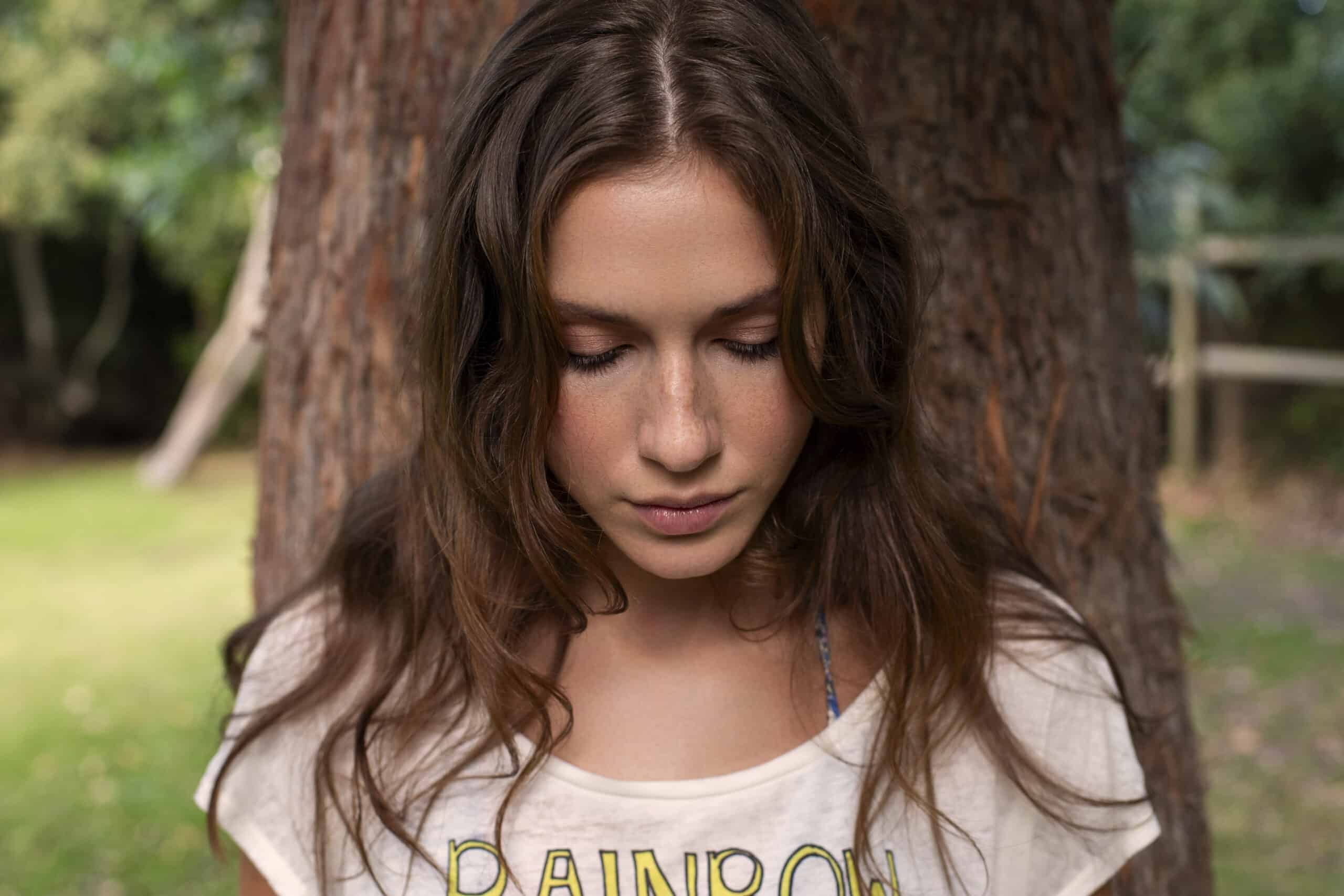 Eine junge Frau mit langen braunen Haaren steht draußen neben einem Baum und blickt nach unten. Sie trägt ein helles T-Shirt mit der Aufschrift „RAINBOW“ und scheint sich in einer friedlichen, natürlichen Umgebung mit viel Grün im Hintergrund zu befinden. © Fotografie Tomas Rodriguez