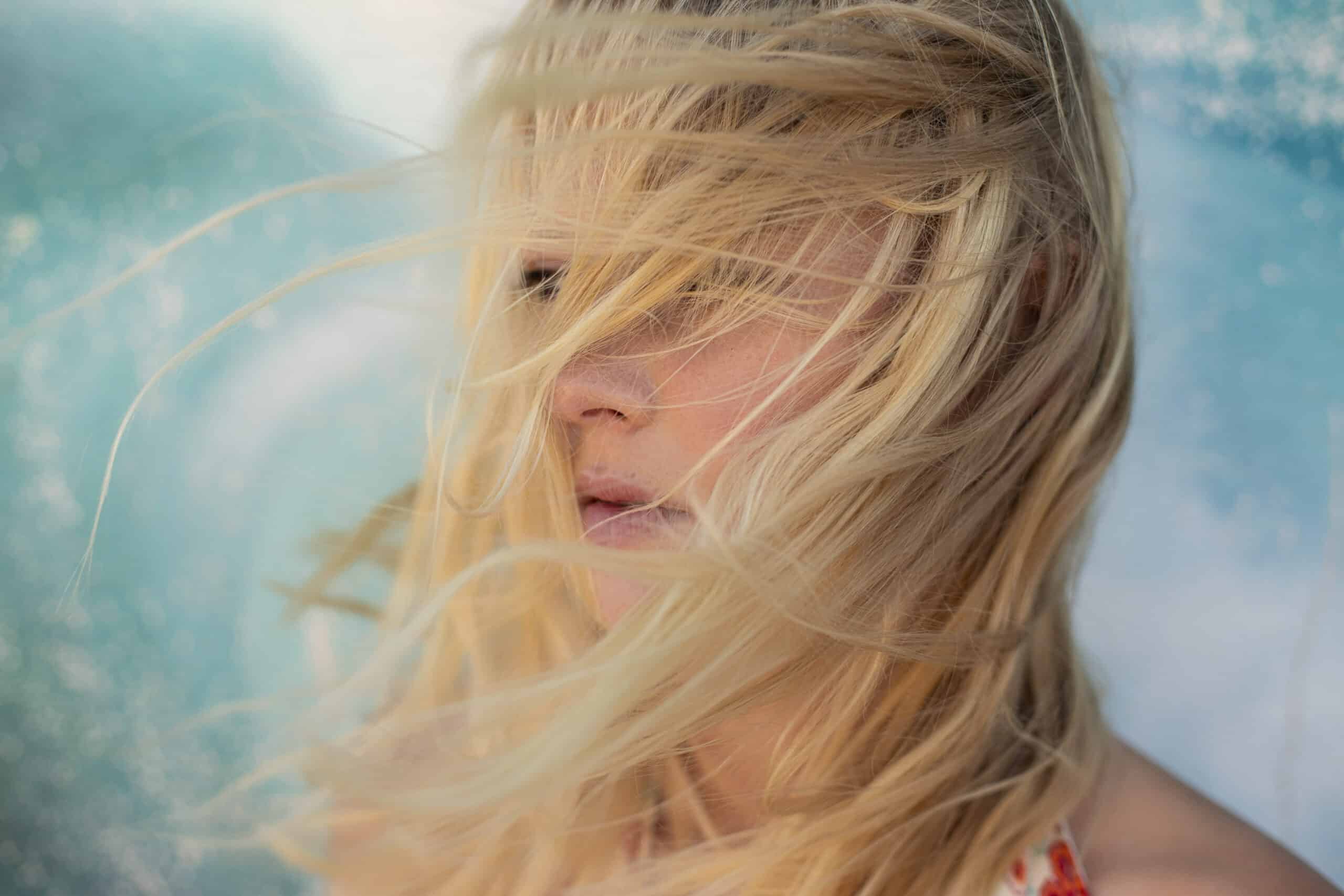 Eine Nahaufnahme einer Person mit langen blonden Haaren, die im Wind wehen und ihr Gesicht teilweise bedecken. Der Hintergrund hat eine verschwommene blaugrüne Farbe, die eine ruhige Umgebung im Freien suggeriert. © Fotografie Tomas Rodriguez