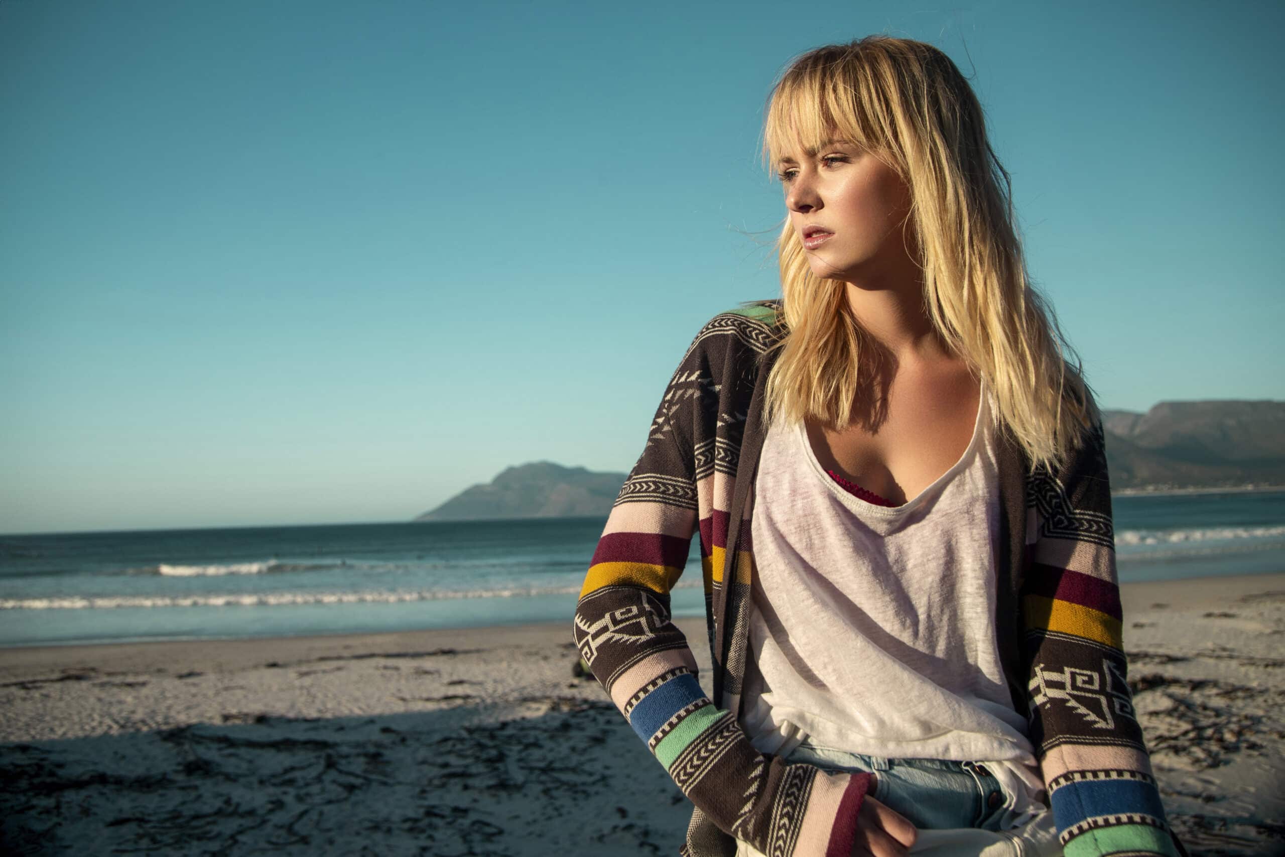 Eine Frau mit langen blonden Haaren steht an einem Strand und blickt in die Ferne. Sie trägt ein lässiges weißes T-Shirt und eine bunt gemusterte Strickjacke. Im Hintergrund sind das Meer und eine Bergkette unter einem klaren blauen Himmel zu sehen. © Fotografie Tomas Rodriguez