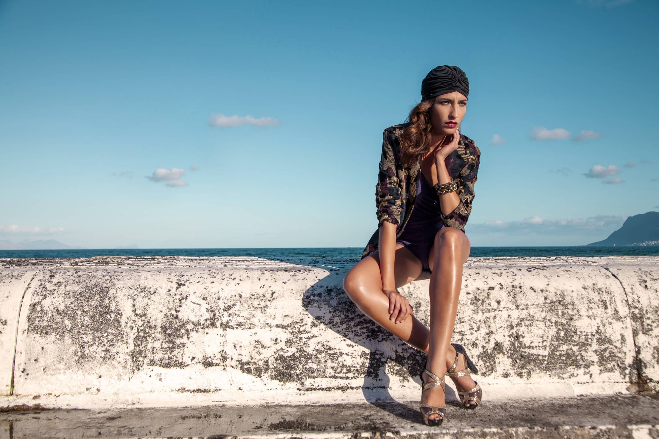 Eine Frau in Tarnjacke, dunklem Kopftuch und Sandalen sitzt mit nachdenklichem Gesichtsausdruck auf einem Betonvorsprung am Meer. Der blaue Himmel und das ruhige Meer bilden einen heiteren Hintergrund. © Fotografie Tomas Rodriguez