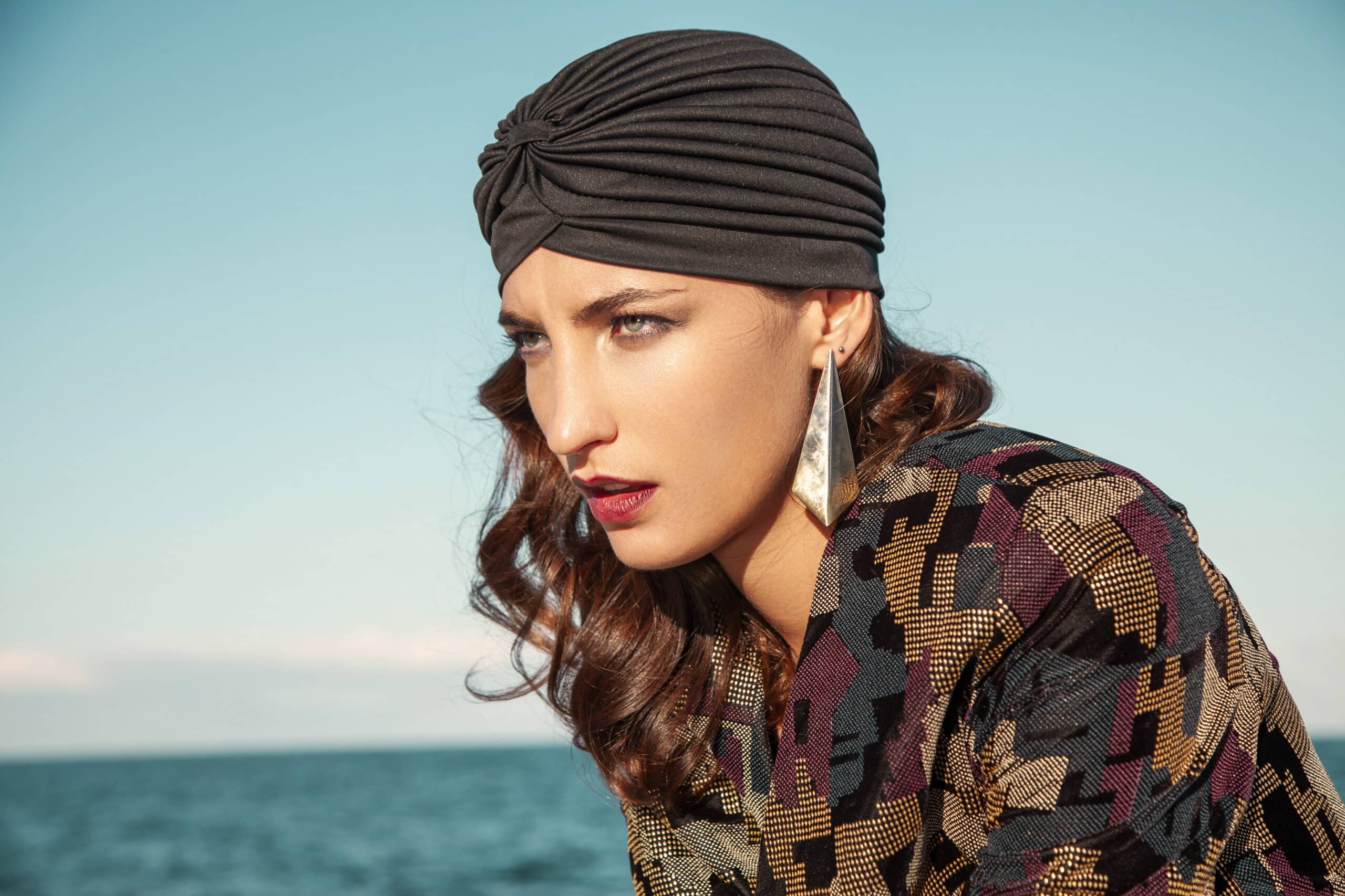 Eine Frau mit welligem braunem Haar, die einen schwarzen Turban und eine Jacke mit geometrischem Muster trägt, blickt nachdenklich in die Ferne. Sie trägt große silberne Ohrringe. Im Hintergrund sind ein ruhiges blaues Meer und ein klarer Himmel zu sehen. © Fotografie Tomas Rodriguez