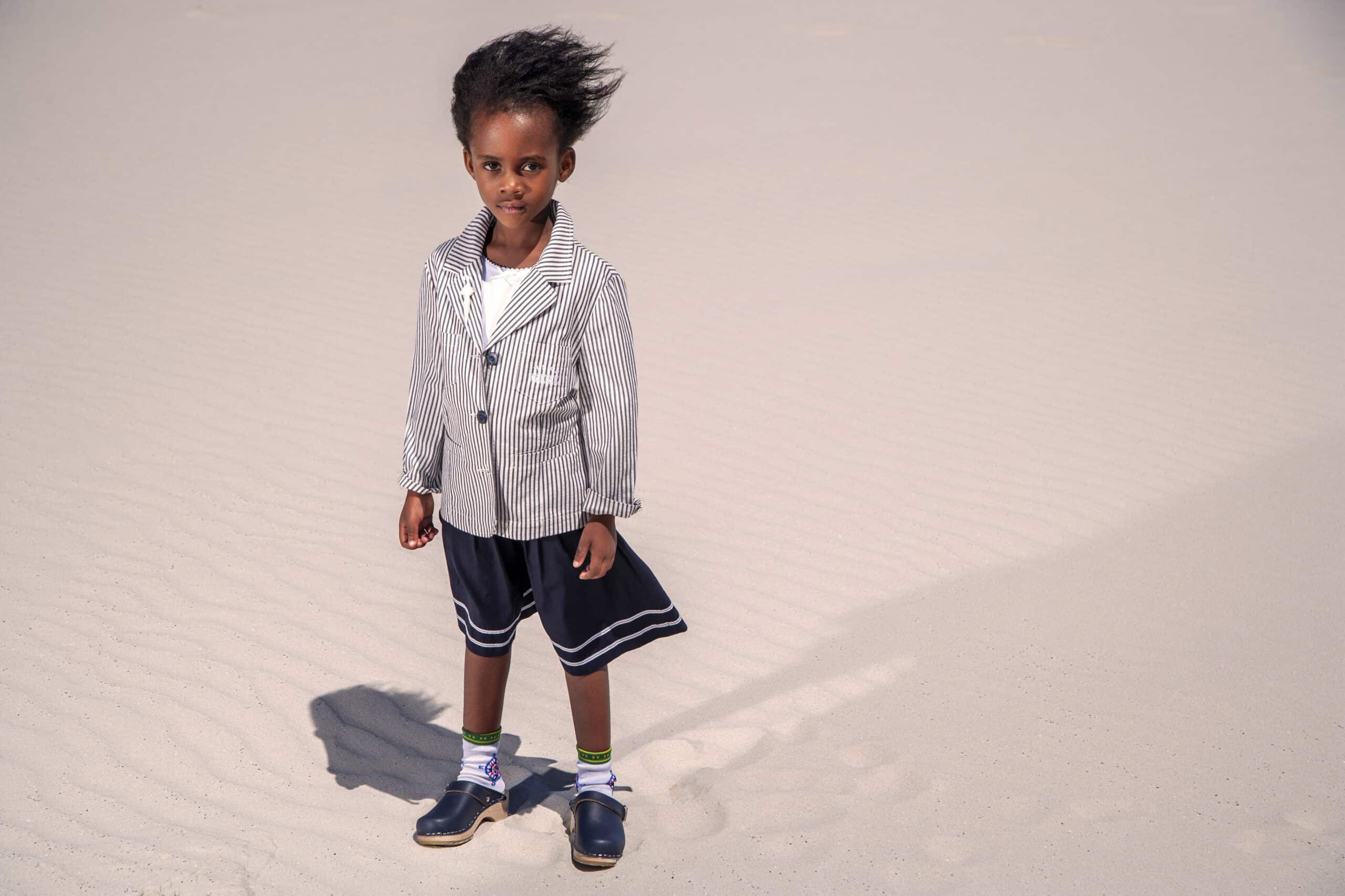Ein kleines Kind steht an einem Sandstrand. Es trägt einen gestreiften Blazer über einem Hemd, marineblaue Shorts und schwarze Schuhe mit weißen Socken. Der Wind weht die Haare des Kindes zur Seite. Der Schatten der Gestalt des Kindes ist im Sand zu sehen. © Fotografie Tomas Rodriguez