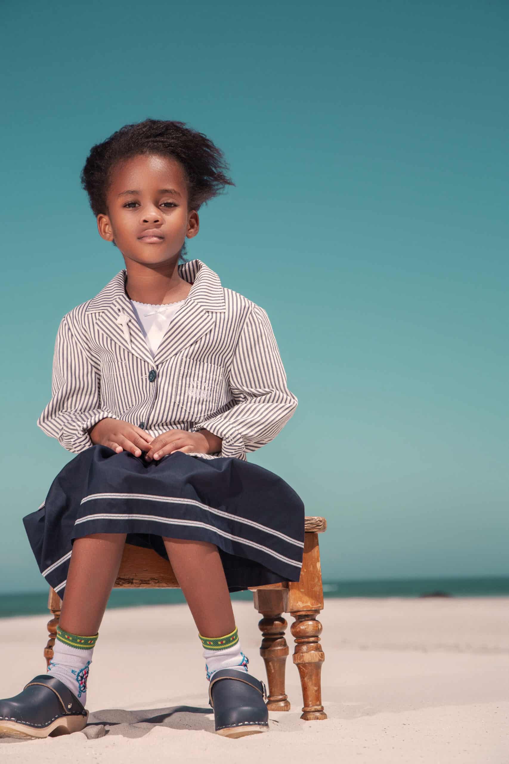 Ein kleines Kind sitzt auf einem kleinen Holztisch am Strand. Das Kind trägt eine gestreifte Jacke, einen marineblauen Rock mit weißem Besatz, bunte Socken und schwarze Schuhe. Im Hintergrund sind ein klarer blauer Himmel und Sand zu sehen. © Fotografie Tomas Rodriguez
