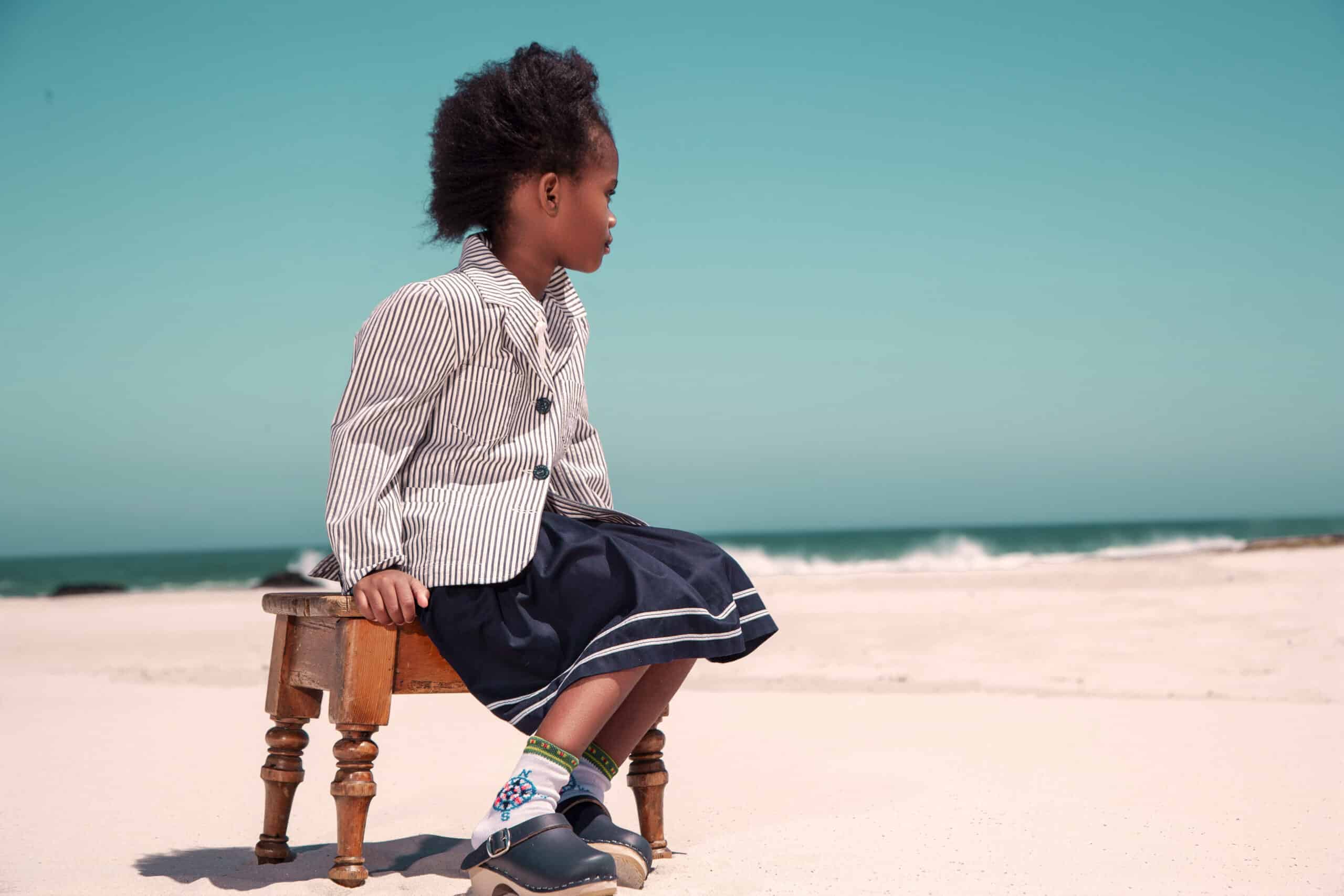 Ein kleines Kind sitzt auf einem kleinen Holzhocker an einem Sandstrand mit Blick auf das Meer. Das Kind trägt eine gestreifte Jacke, einen marineblauen Rock mit weißem Besatz und marineblaue Schuhe. Der Himmel ist klar und blau und verschmilzt mit dem Meer im Hintergrund. © Fotografie Tomas Rodriguez