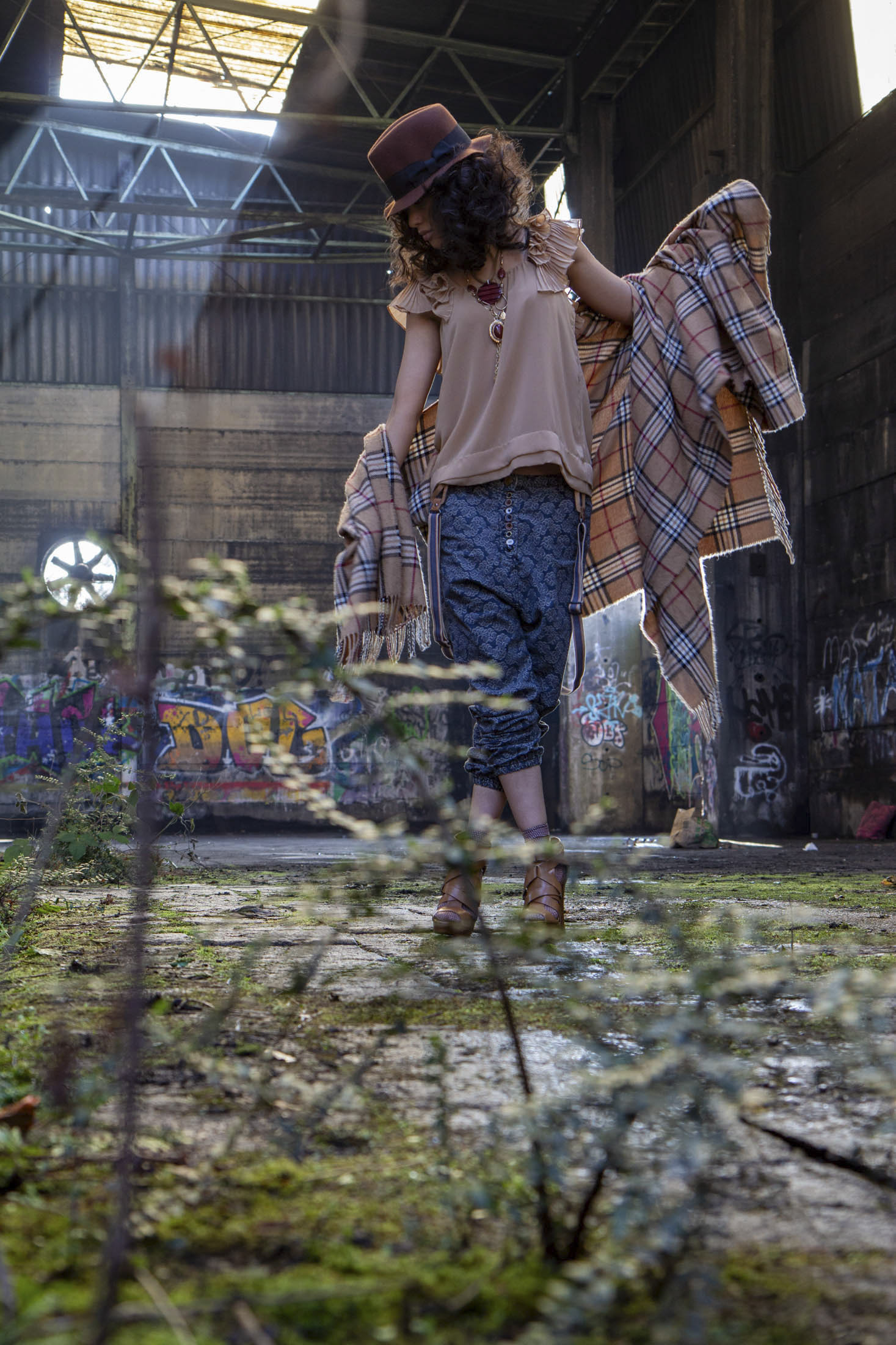Eine Person mit lockigem Haar, einem braunen Hut und schicker Freizeitkleidung steht in einem verlassenen, mit Graffiti bedeckten Gebäude mit einem großen Fenster. Sie hält einen karierten Schal in der Hand und der Boden ist mit Grünpflanzen überwuchert, was eine künstlerische, urbane Verfallsszenerie schafft. © Fotografie Tomas Rodriguez