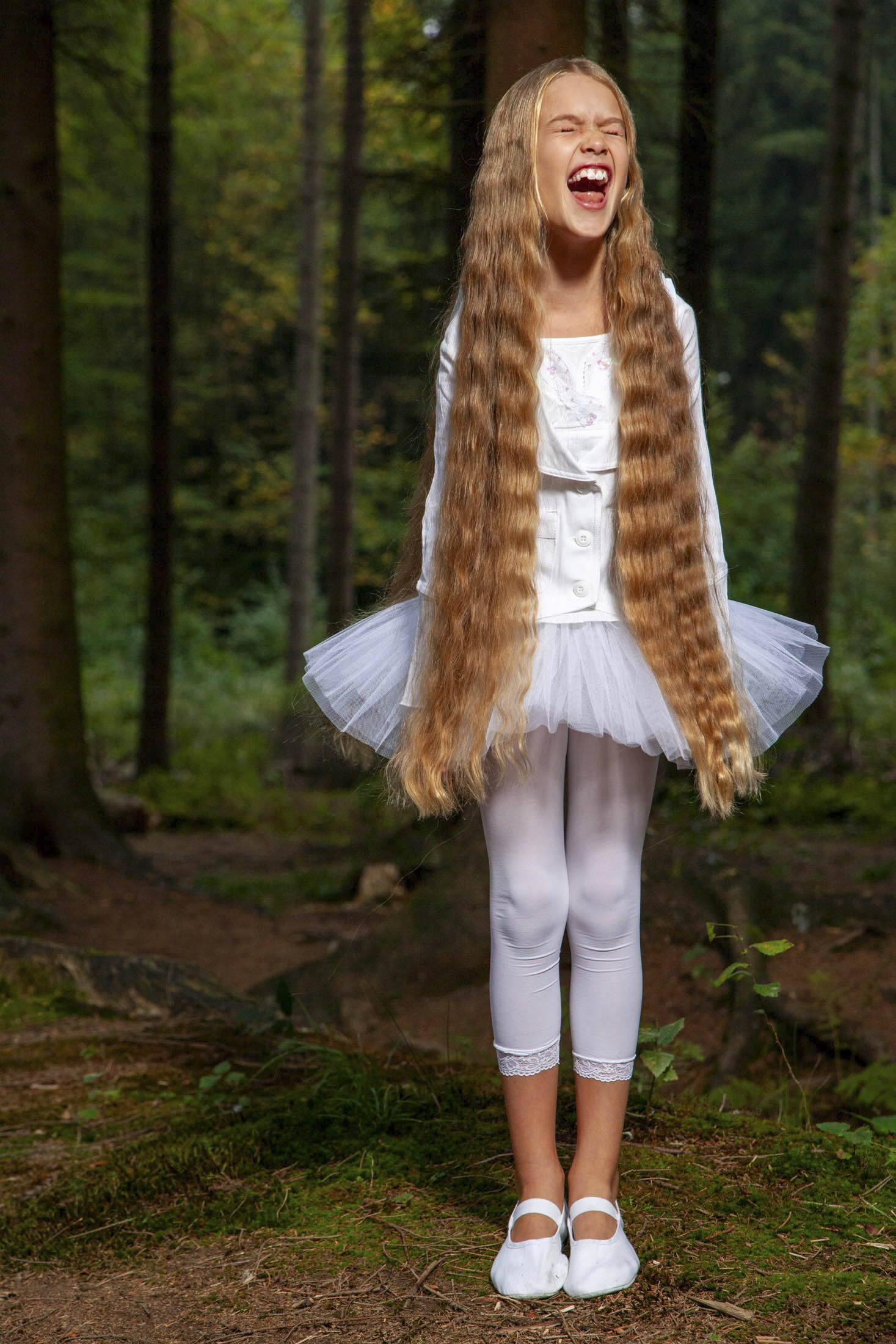 Ein junges Mädchen mit sehr langem, welligem Haar lacht laut in einem Wald. Sie trägt ein weißes Outfit bestehend aus einem Tutu, einem langärmligen Oberteil, Leggings und Ballettschuhen. Im Hintergrund sind hohe Bäume zu sehen und der Boden ist mit Grünpflanzen und Laub bedeckt. © Fotografie Tomas Rodriguez