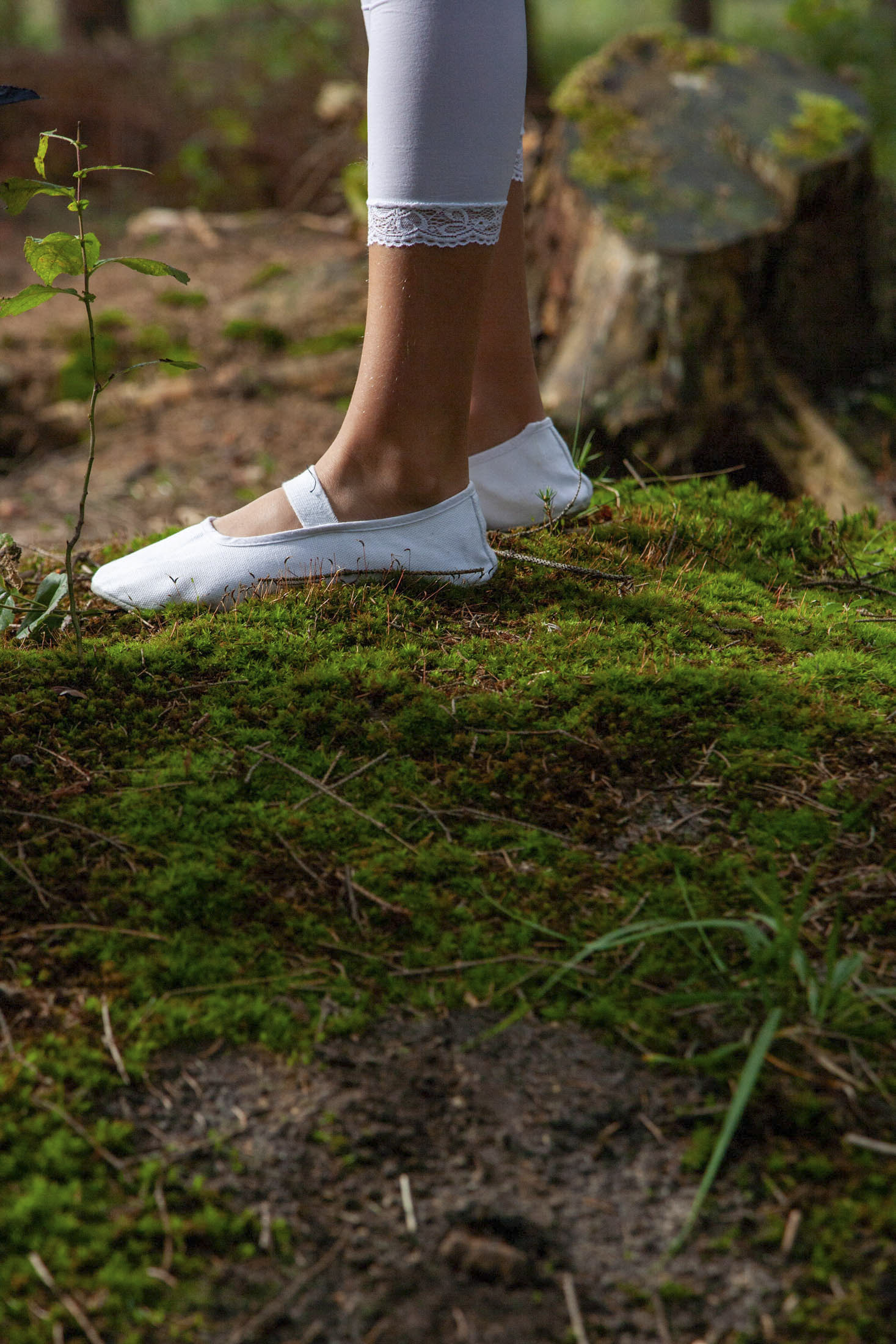 Eine Person in weißen Leggings und dazu passenden weißen Ballerinas steht auf einem Moosfleck in einer Waldlandschaft. Das Bild ist von unterhalb des Knies abwärts aufgenommen und zeigt Details des moosigen Bodens und Andeutungen von Baumstümpfen im Hintergrund. © Fotografie Tomas Rodriguez