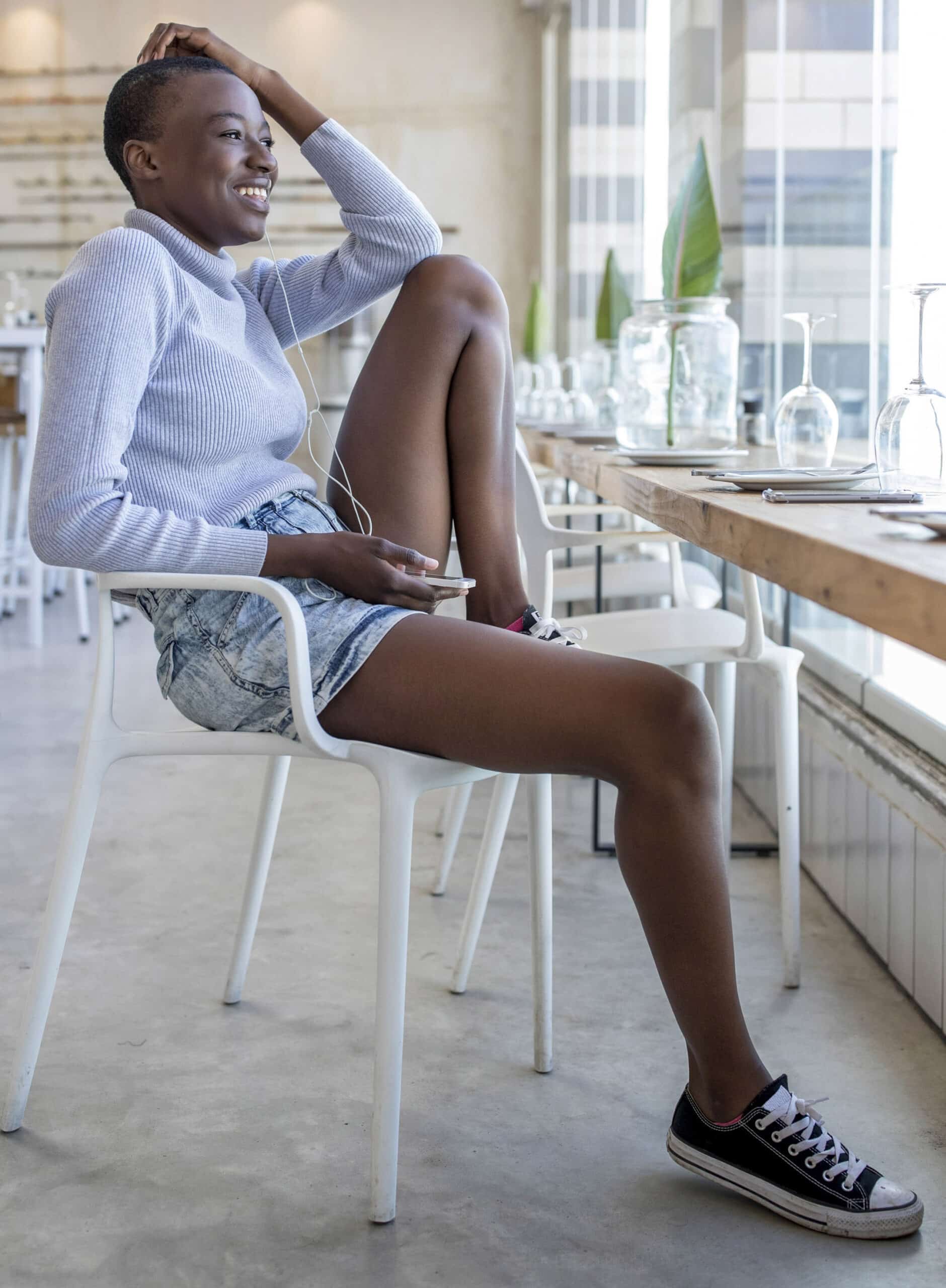 Eine Frau sitzt lässig auf einem weißen Stuhl in einem hellen Café. Sie trägt ein hellblaues Langarmshirt, Jeansshorts und schwarze Turnschuhe. Sie lächelt und hat einen Arm auf dem Kopf, trägt Kopfhörer und hält ein Telefon in der Hand. Auf dem Tisch neben ihr stehen mehrere Glasflaschen. © Fotografie Tomas Rodriguez