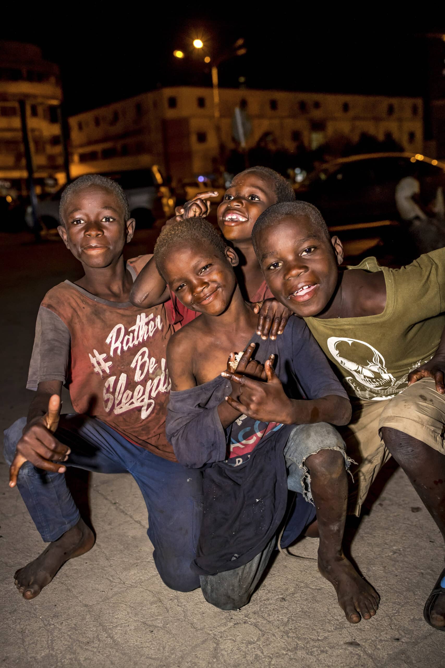 Vier Jungen posieren nachts in einer städtischen Umgebung für ein Foto. Sie lächeln und gestikulieren mit den Händen. Sie tragen legere Kleidung mit Abnutzungs- und Schmutzspuren. Im Hintergrund sind Gebäude und Straßenlaternen zu sehen, die zur geschäftigen Abendatmosphäre beitragen. © Fotografie Tomas Rodriguez