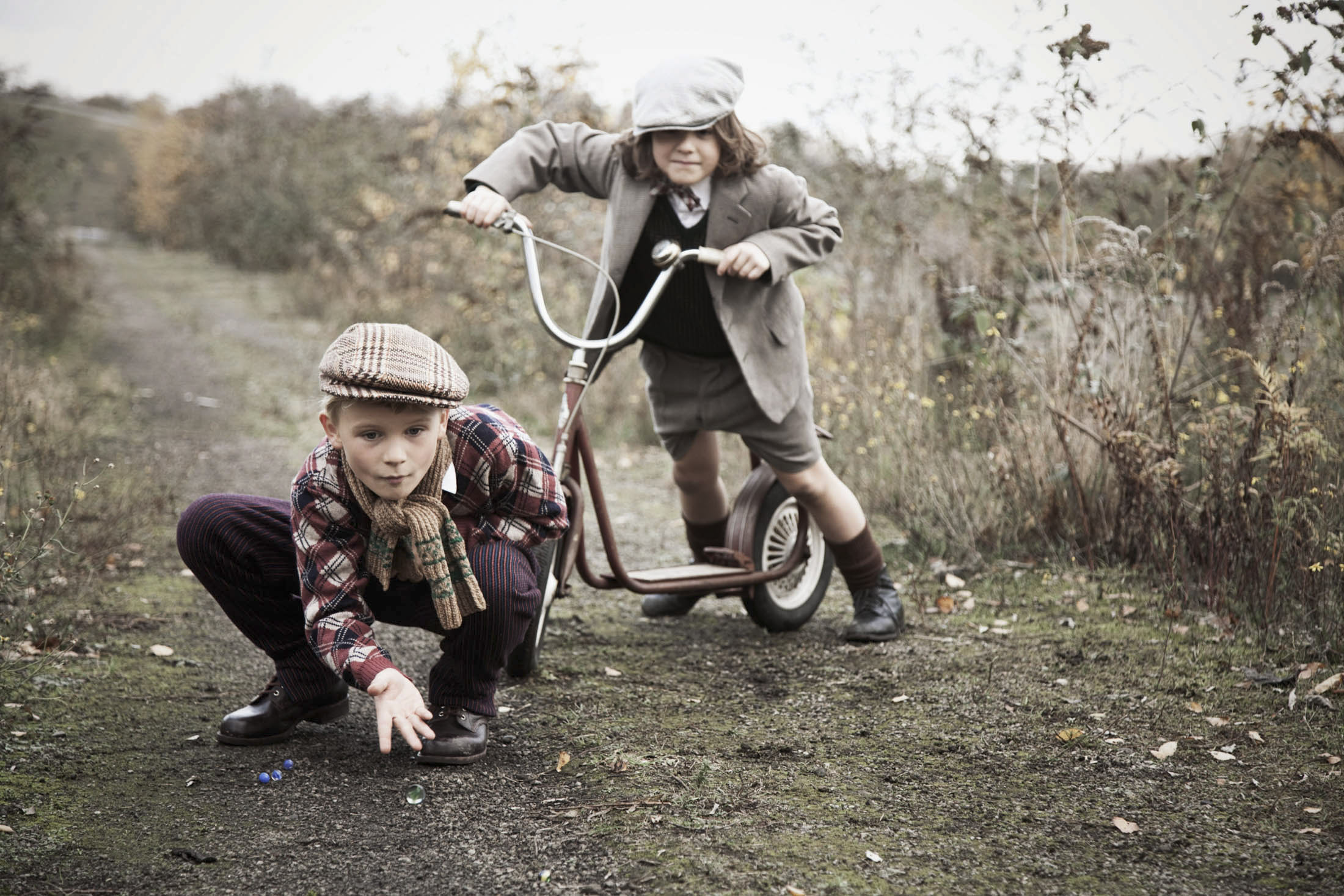 Zwei Kinder in Vintage-Kleidung spielen auf einem Kiesweg. Ein Kind hockt und konzentriert sich auf Murmeln, während das andere hinter einem klassischen roten Dreirad steht und aussieht, als wäre es bereit loszufahren. Der Hintergrund ist eine natürliche, ländliche Umgebung mit überwucherter Vegetation auf beiden Seiten. © Fotografie Tomas Rodriguez