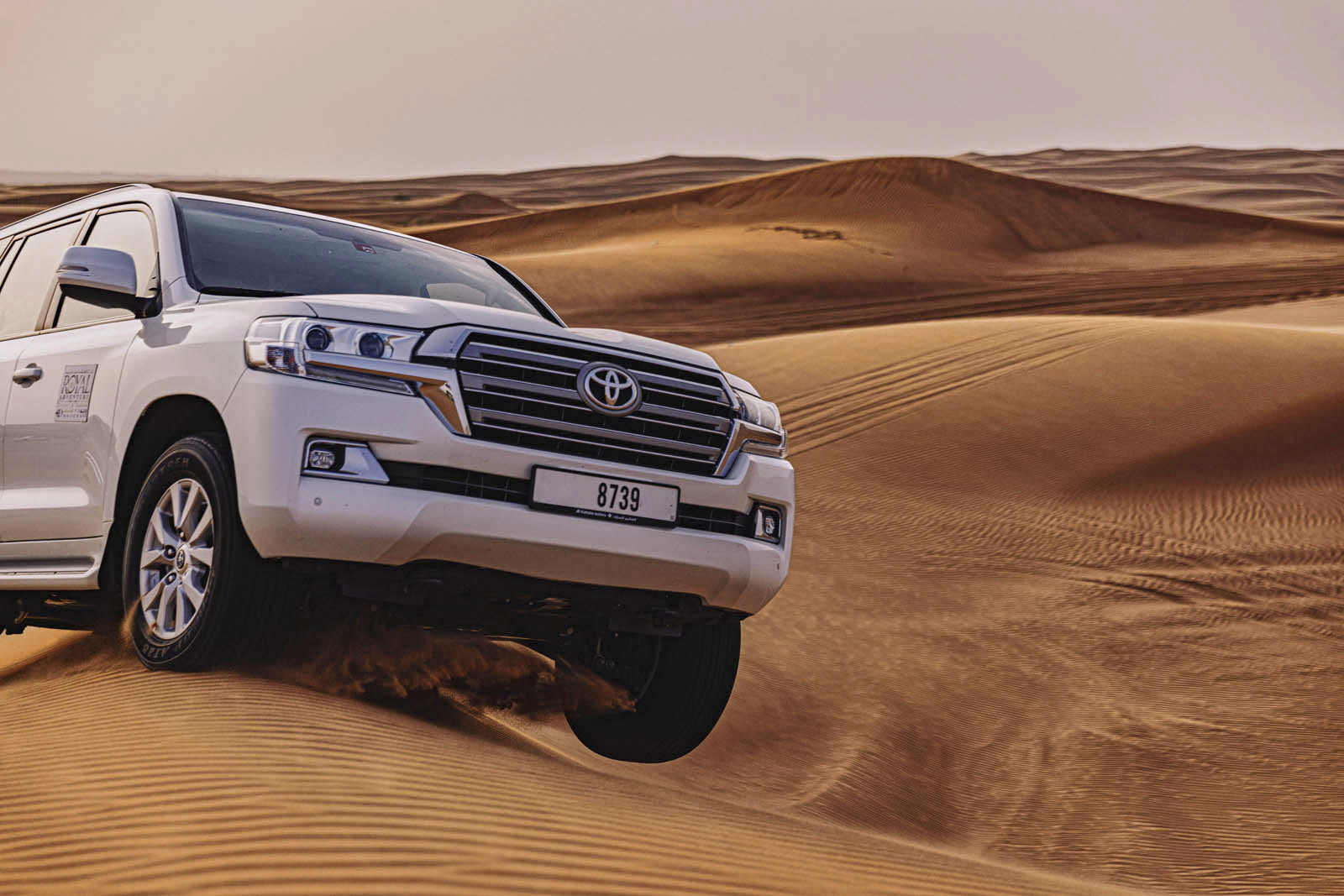 Ein weißer Toyota SUV fährt auf Sanddünen in einer Wüstenlandschaft. Das Fahrzeug navigiert leicht geneigt durch das unebene Gelände mit großen Sanddünen im Hintergrund unter einem klaren Himmel. Das Nummernschild auf der Vorderseite des SUV lautet „8739“. © Fotografie Tomas Rodriguez