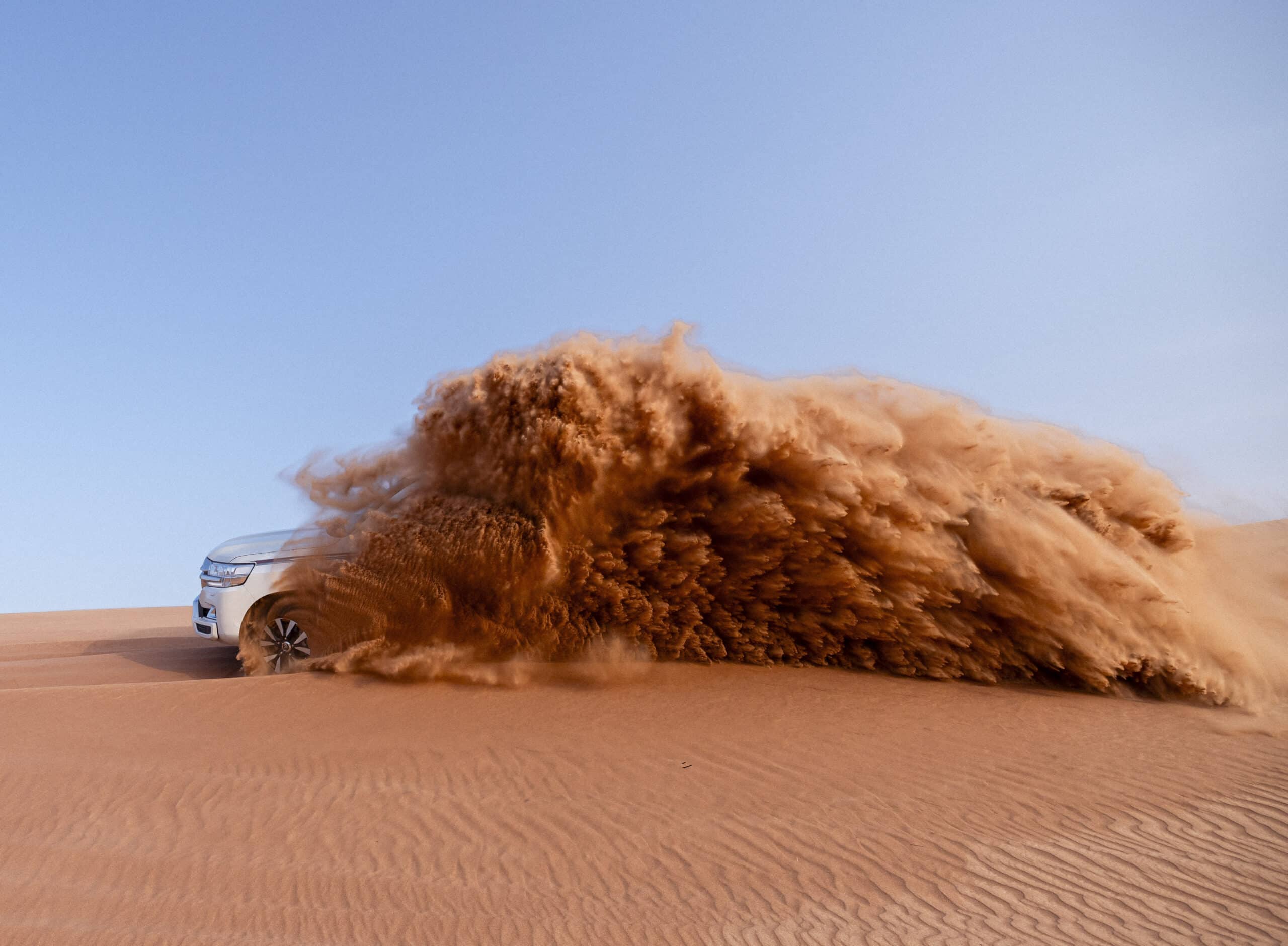Ein Fahrzeug fährt durch eine Wüste und erzeugt dabei eine große Sandwolke. Seine Reifen sind teilweise im Sand vergraben, was seine Geschwindigkeit und die dynamische Bewegung des Fahrzeugs über das sandige Gelände betont. Der Himmel darüber ist klar und blau. © Fotografie Tomas Rodriguez