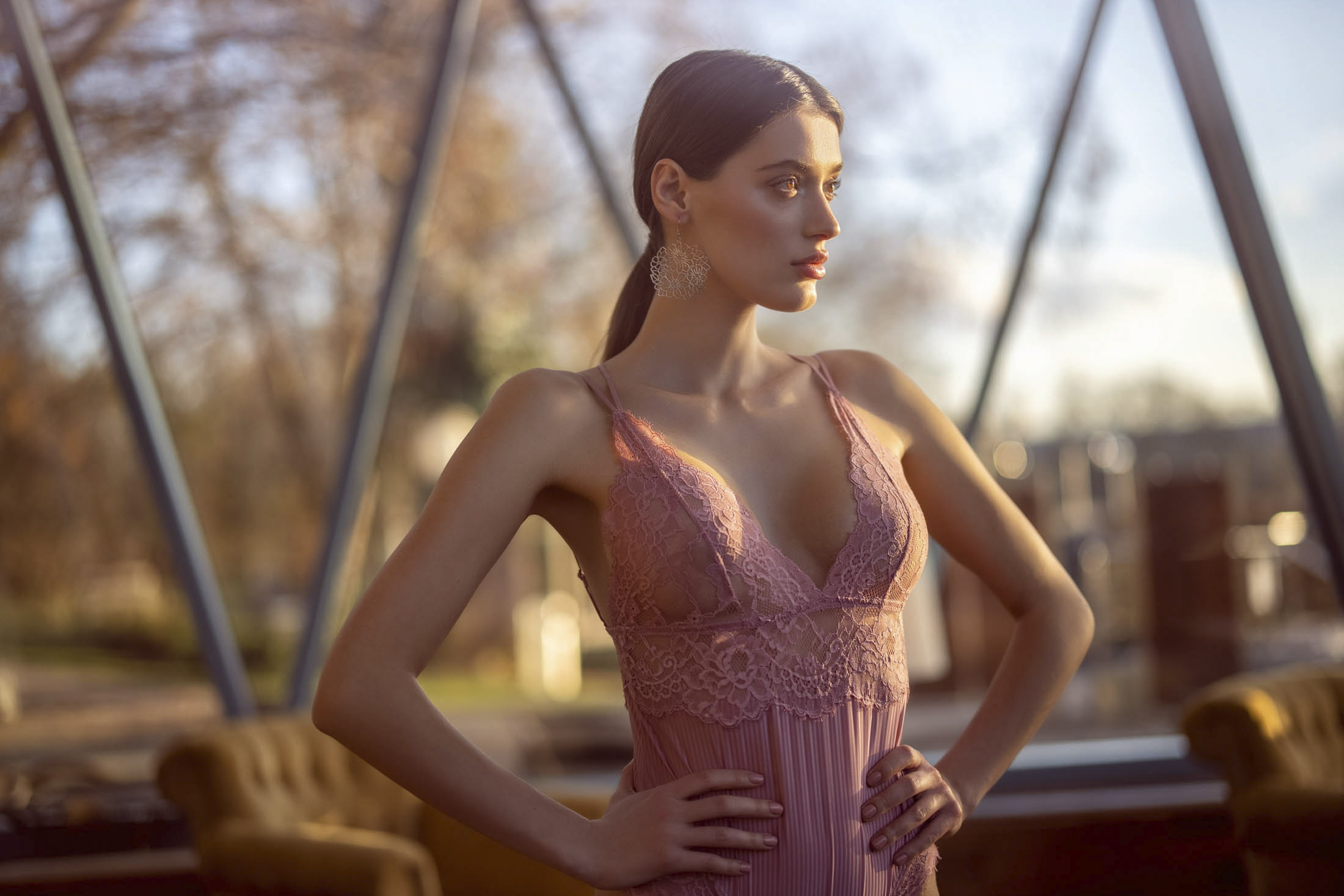Eine Frau in einem rosa Spitzenkleid steht mit den Händen in den Hüften im Haus. Im Hintergrund sind große dreieckige Fenster zu sehen, durch die man eine Außenszene mit Bäumen und einem Gebäude sehen kann. Die Beleuchtung erzeugt einen weichen, warmen Schein auf ihr. © Fotografie Tomas Rodriguez