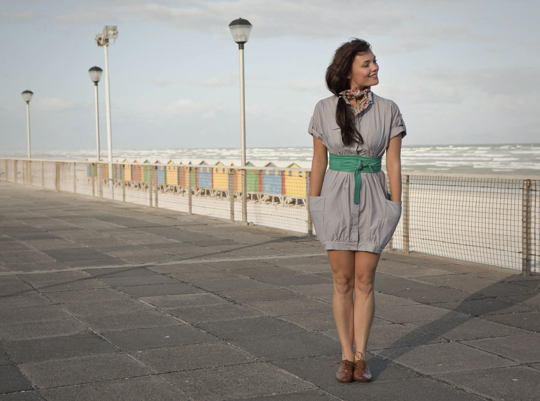 Eine Frau in einem grauen Kleid mit grünem Besatz steht auf einer gepflasterten Promenade in der Nähe eines Strandes. Sie hat die Hände in den Taschen ihres Kleides und blickt lächelnd nach links. Hinter ihr sind bunte Strandhütten und ein paar Laternenpfähle zu sehen, im Hintergrund die Meereswellen. © Fotografie Tomas Rodriguez