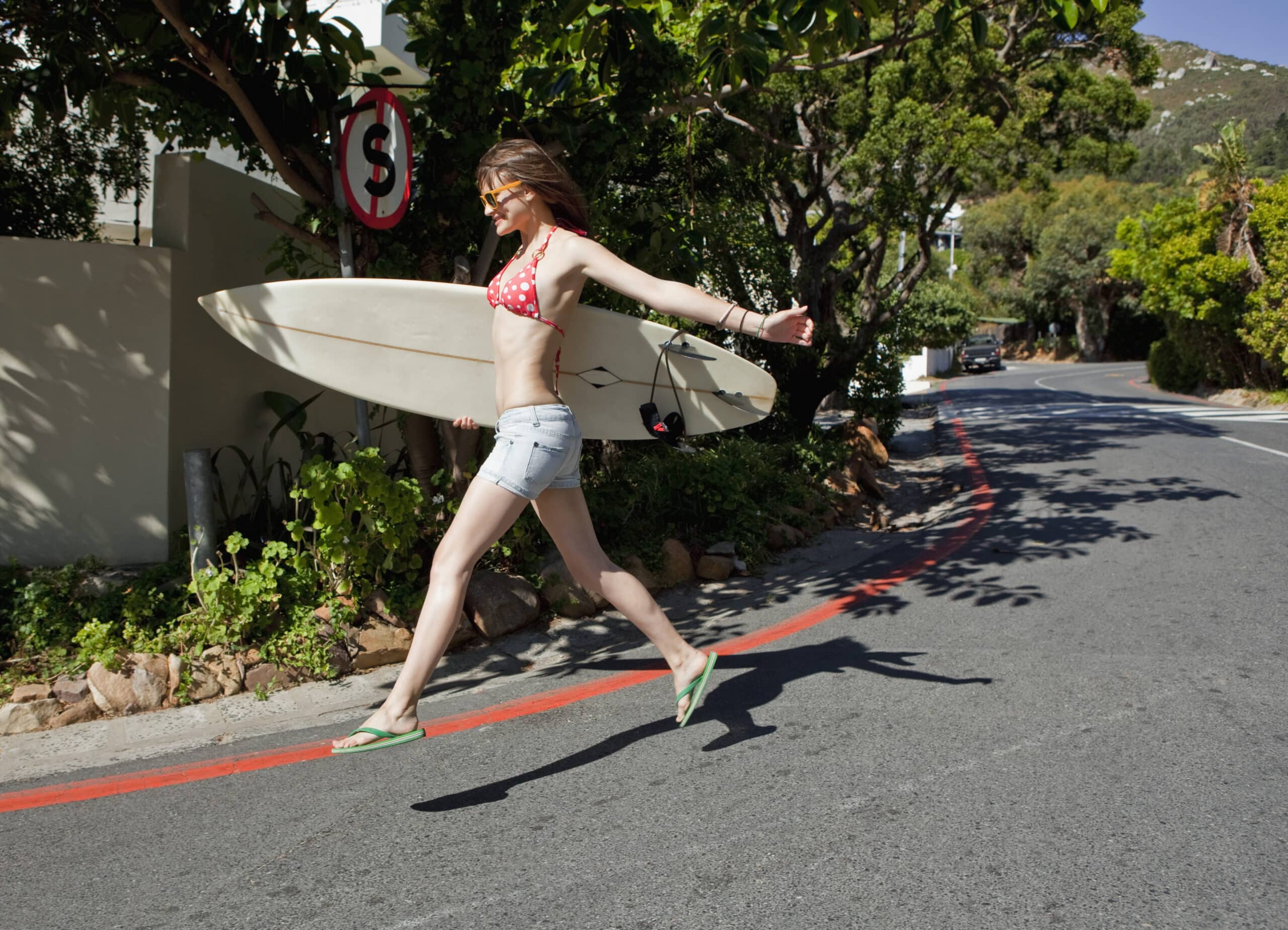 Eine Frau in einem roten Bikinioberteil mit Punkten, Jeansshorts und Flip-Flops geht energisch über eine sonnige Straße. Sie trägt ein Surfbrett unter dem Arm. Im Hintergrund sind Bäume und ein Berg zu sehen, links von ihr steht ein „Einfahrt verboten“-Schild. © Fotografie Tomas Rodriguez